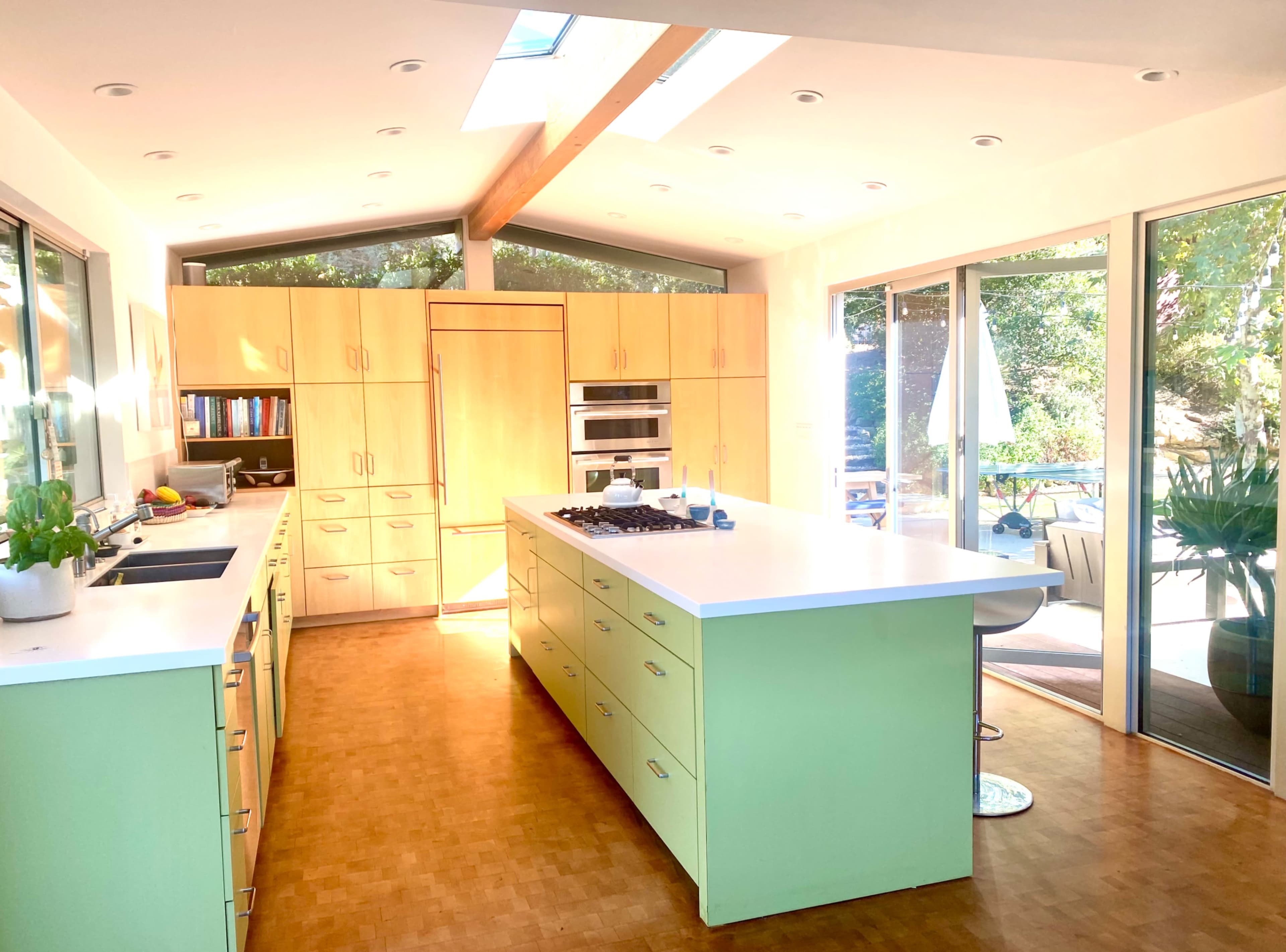 A modern kitchen features light green cabinetry, a large central island, and ample natural light from windows and skylights.