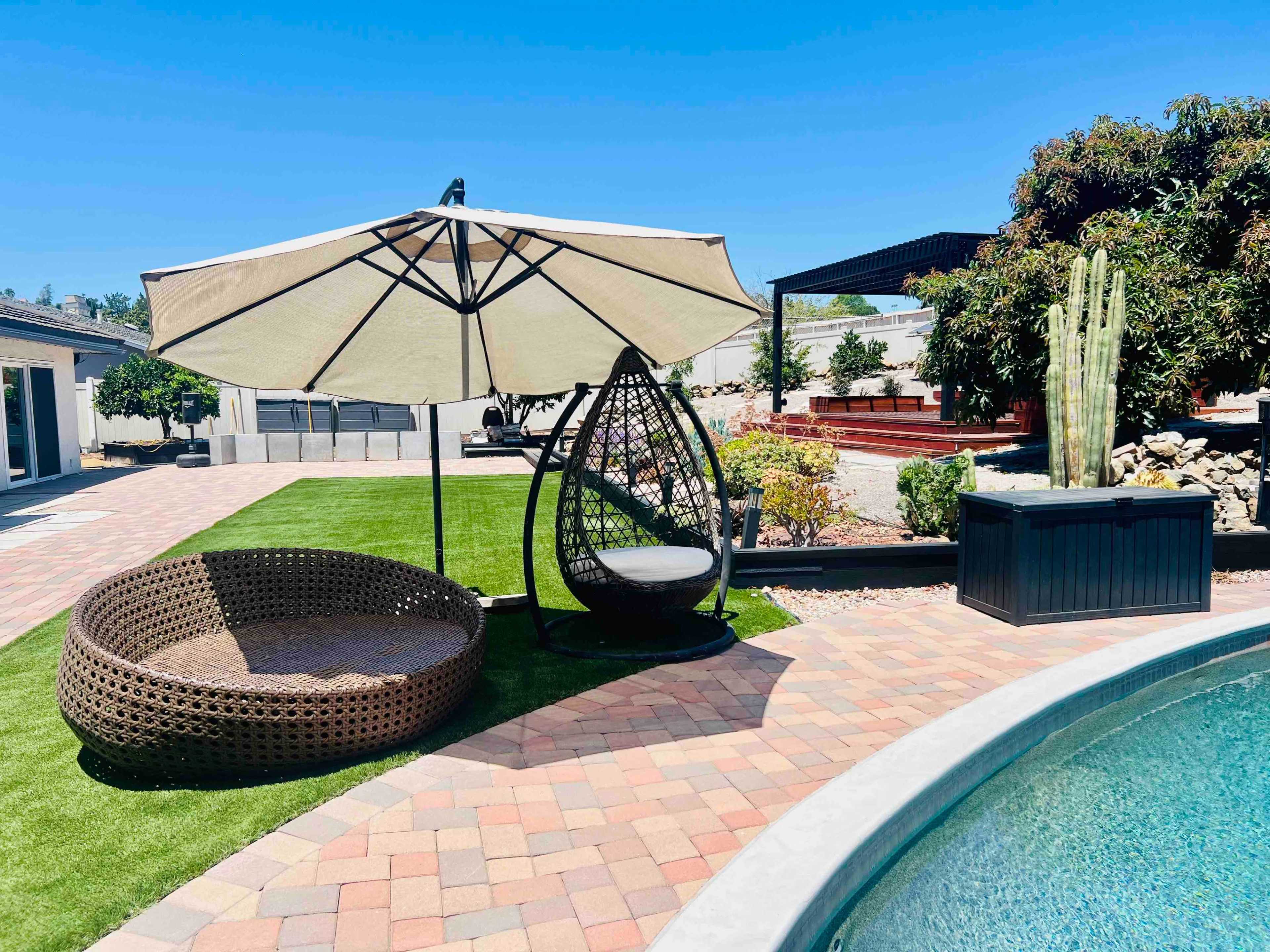 The image shows a well-maintained backyard featuring a swimming pool, a large umbrella, a hanging chair, and a circular wicker sofa surrounded by landscaped areas.