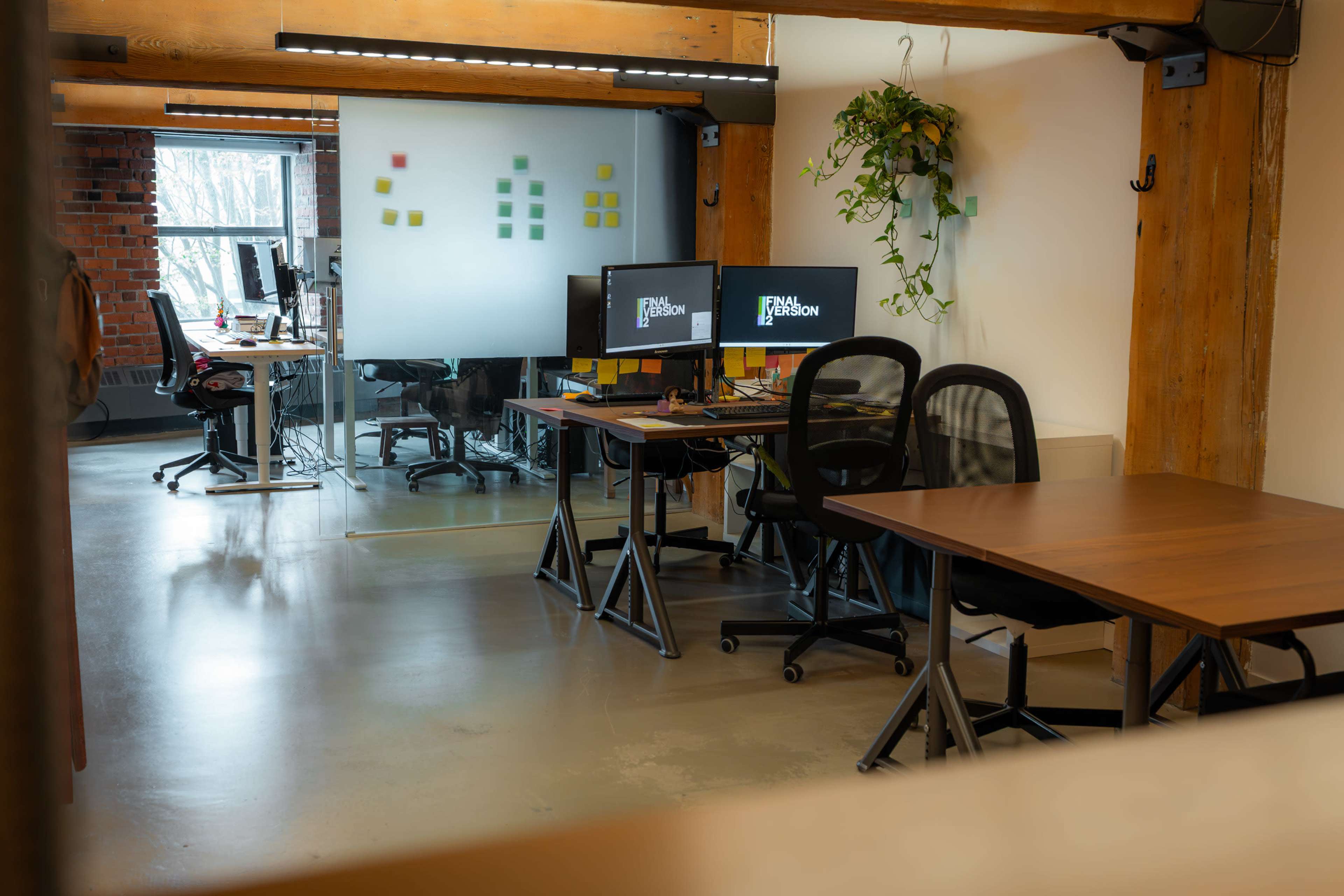 The image shows a modern office space featuring several workstations, a glass wall with sticky notes, and greenery hanging from the ceiling.