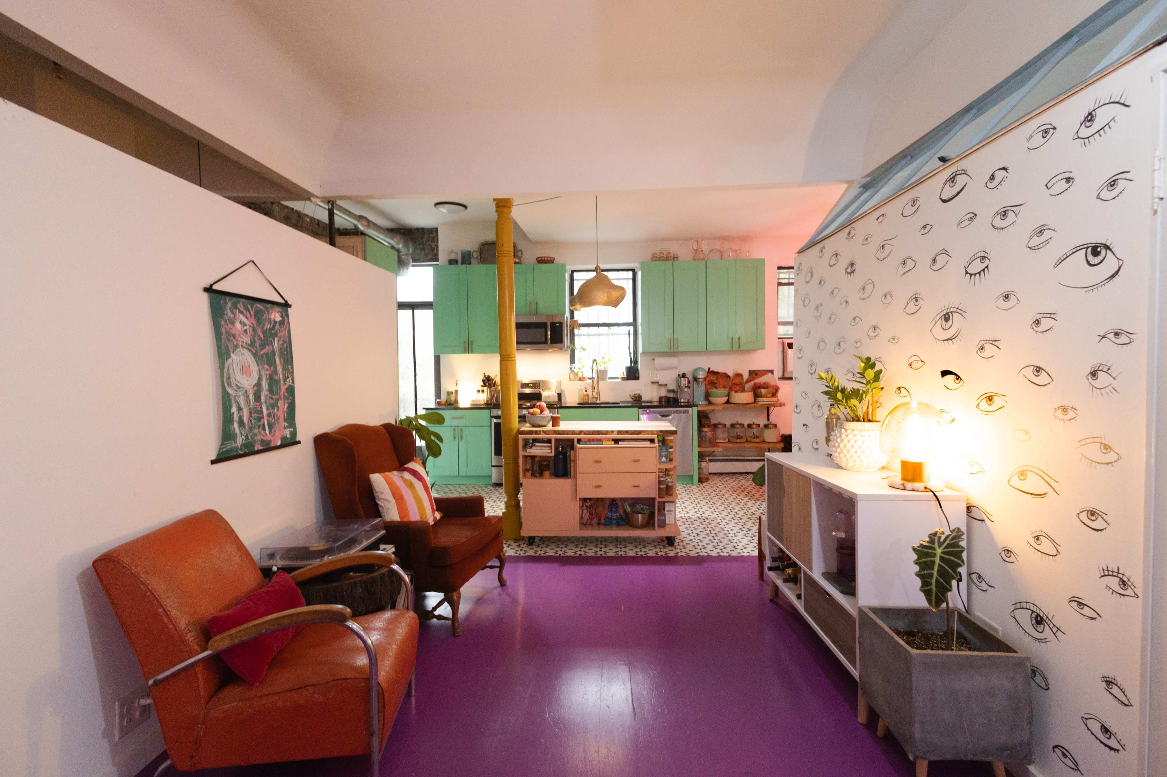 The image shows a modern apartment with a colorful kitchen, featuring mint green cabinets, a purple floor, and an accent wall decorated with eye illustrations.