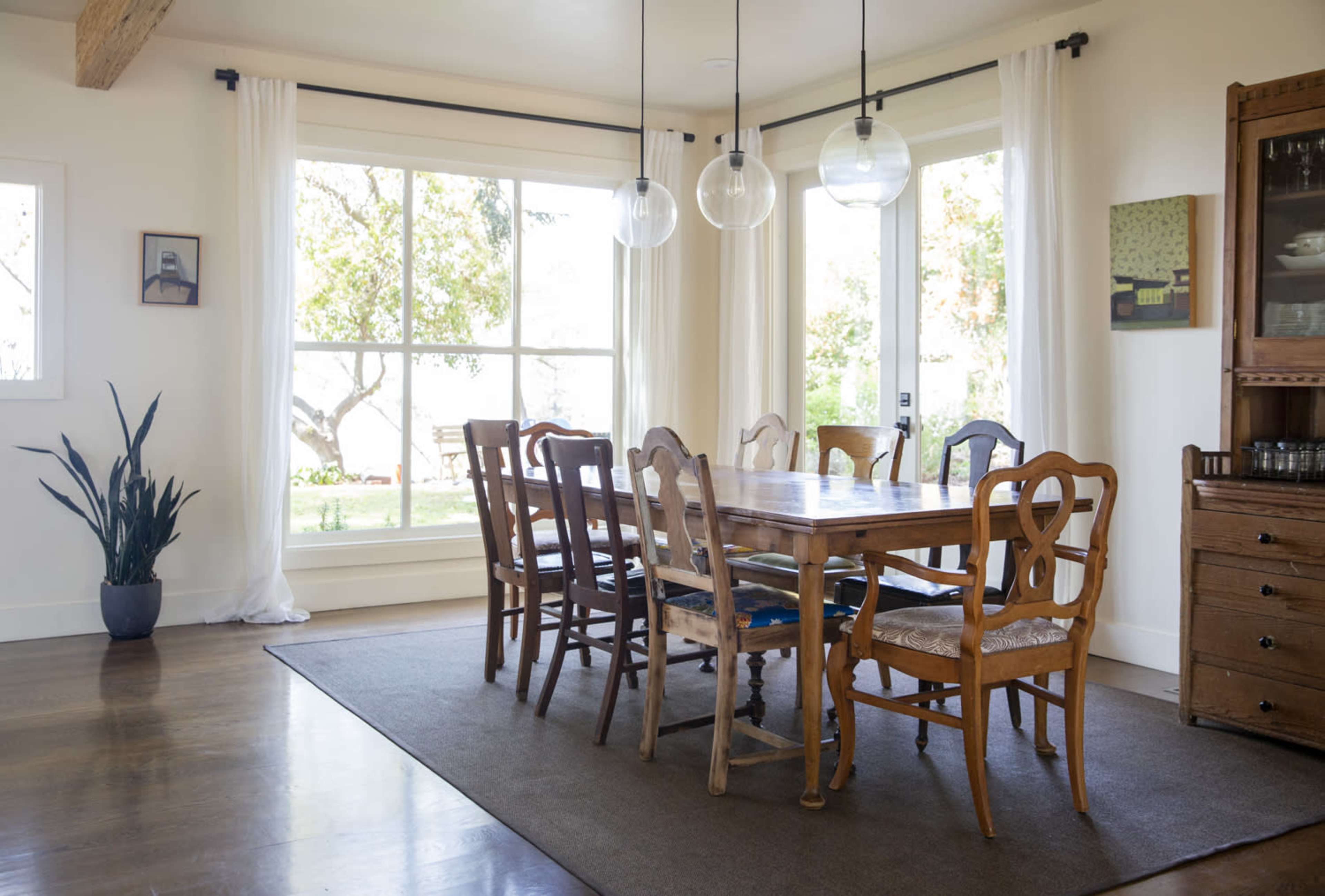 A dining room features a large wooden table surrounded by eight chairs, with large windows letting in natural light and a plant in the corner.
