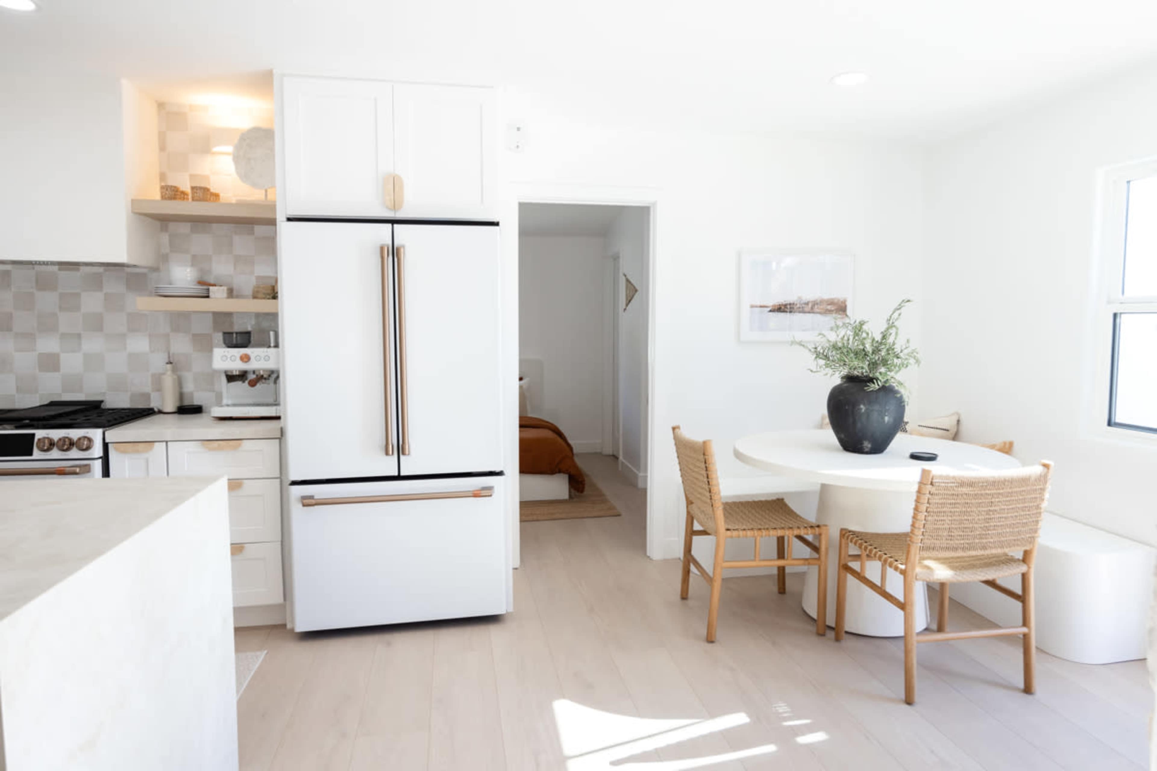 A modern kitchen and dining area features a white refrigerator, a round dining table with woven chairs, and a doorway leading to a bedroom.
