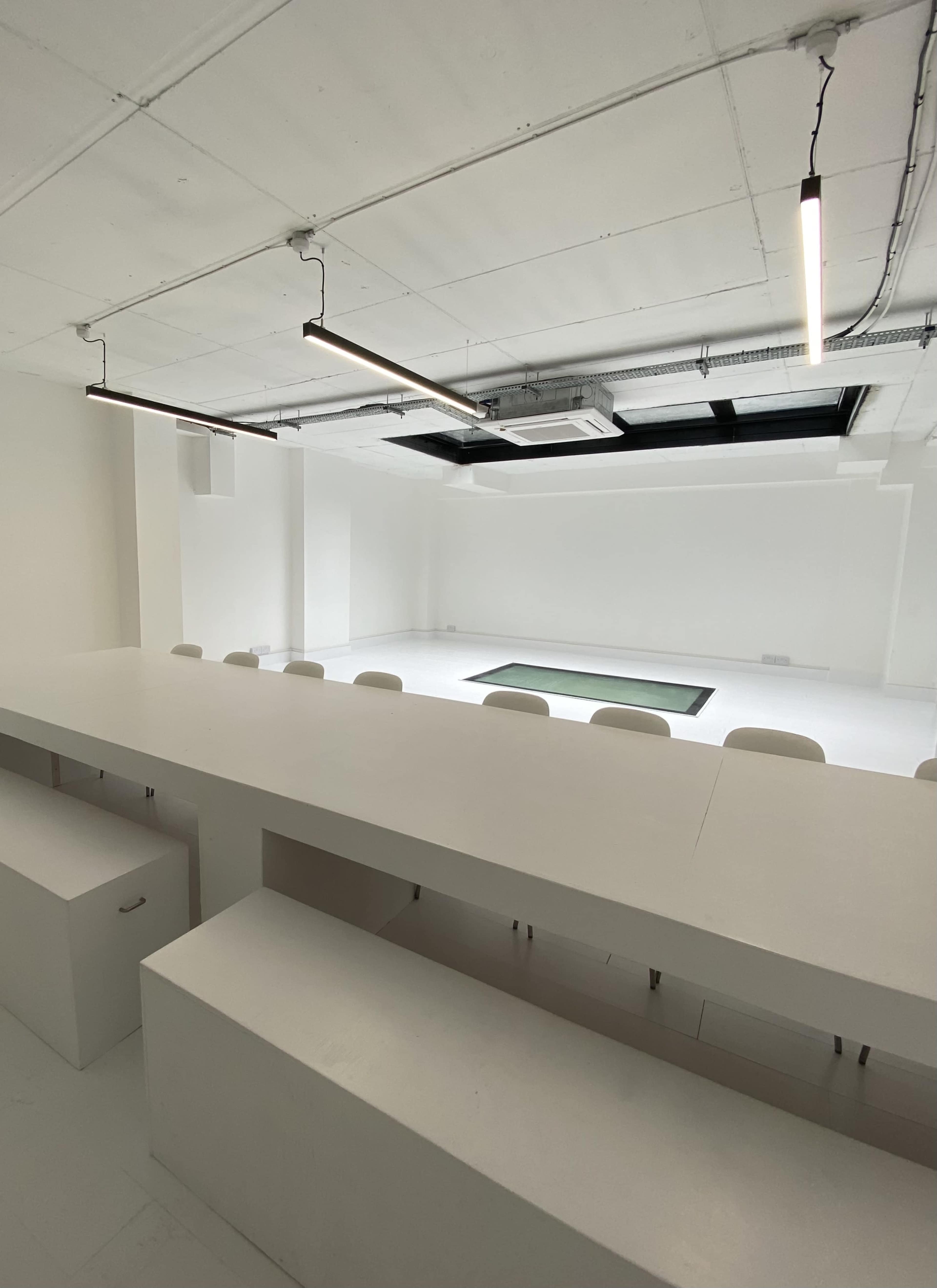 A minimalist, bright room with a long white table and chairs facing an empty space with a green mat on the floor.