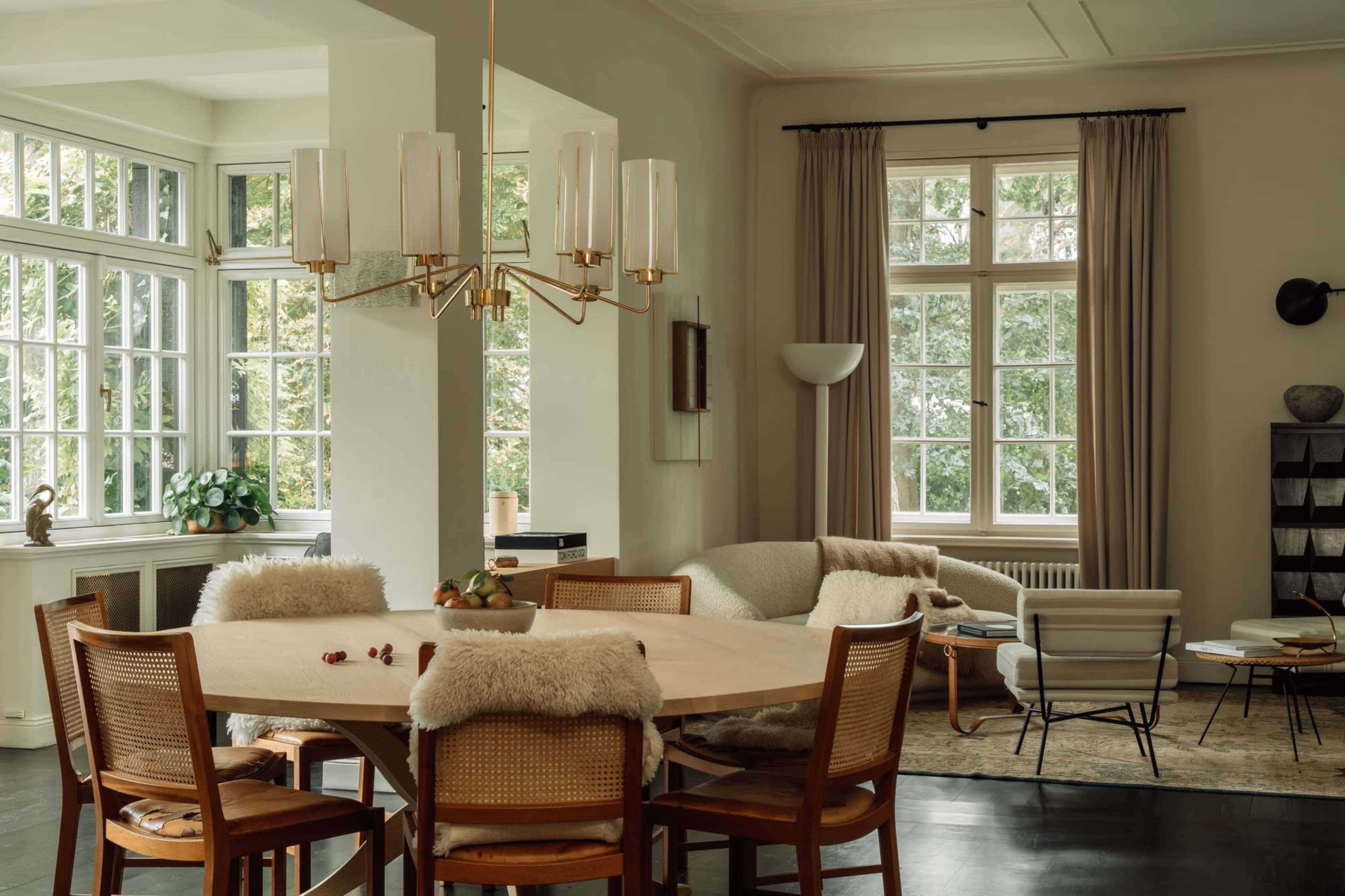 A modern dining area features a round table surrounded by wooden chairs with soft seat covers, large windows letting in natural light, and a chic chandelier overhead.