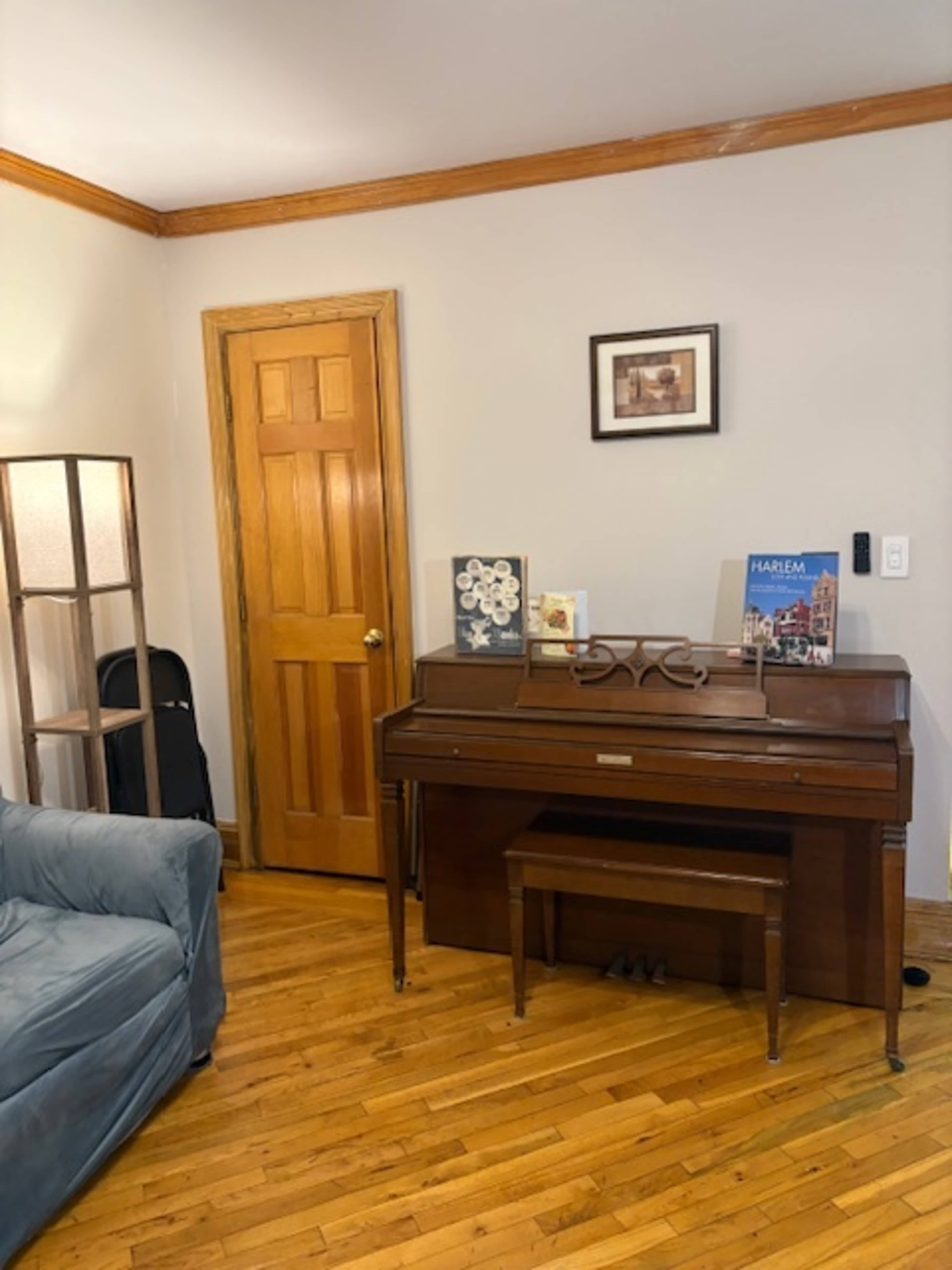 The image shows a wooden piano placed against a wall in a room with hardwood floors, a door on one side, and a lamp nearby.