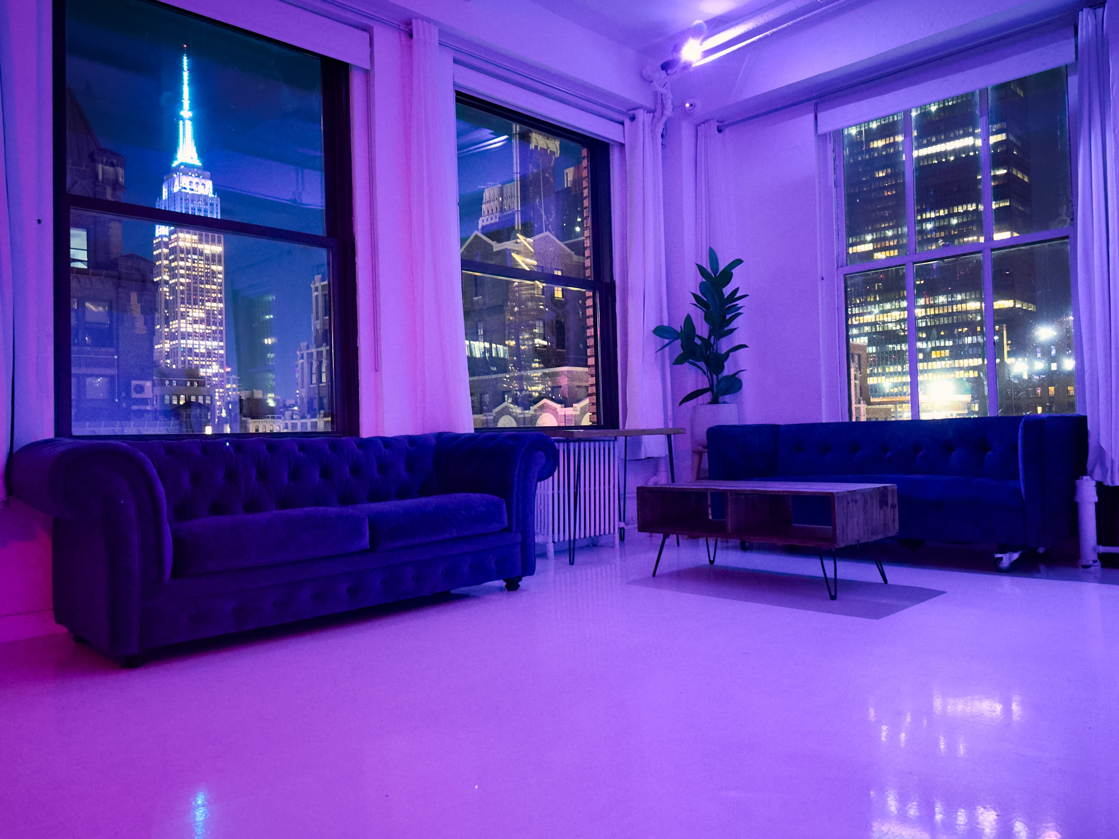 A modern interior with two sofas facing a coffee table, and large windows revealing a city skyline featuring the Empire State Building illuminated at night.