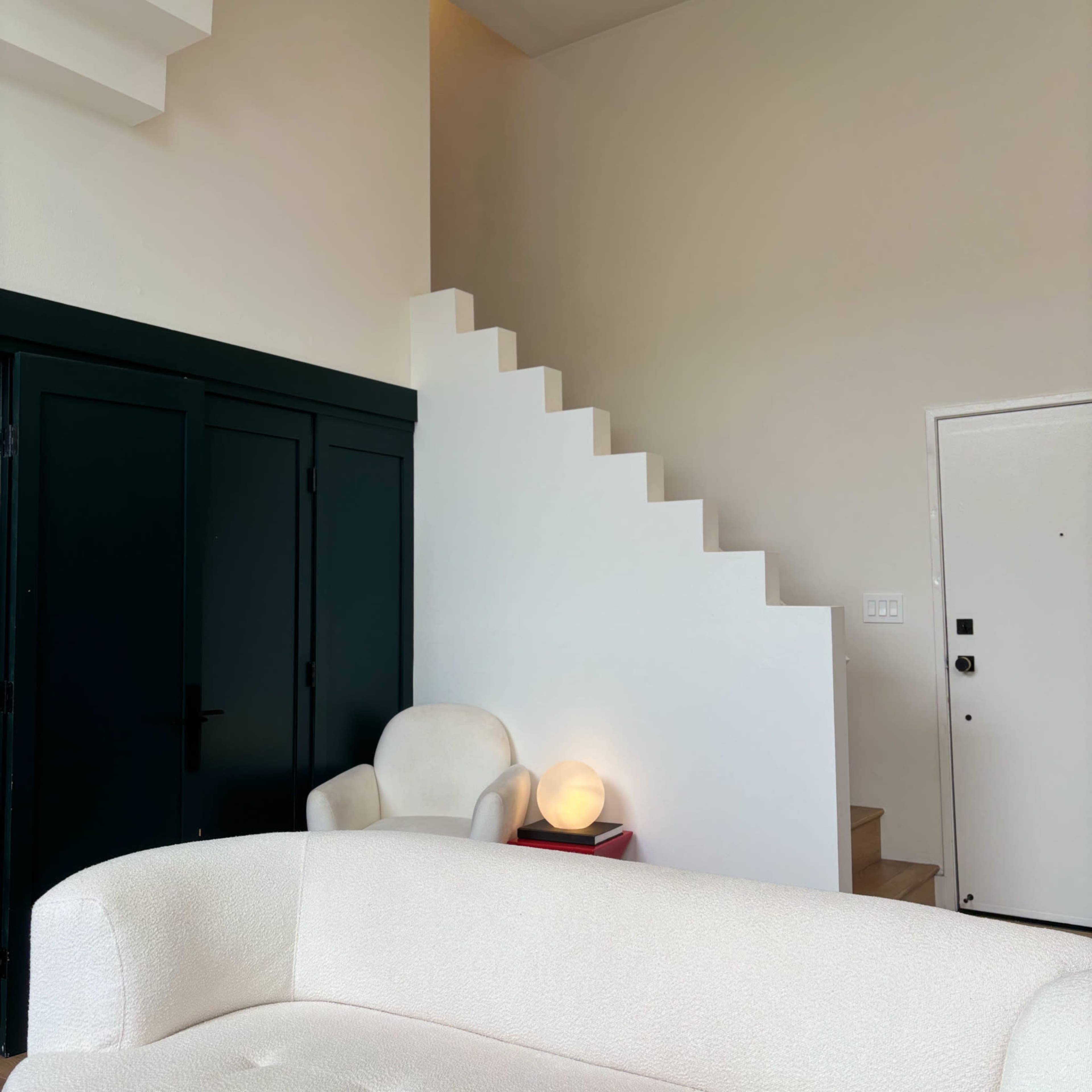 A white couch sits in a modern room with a staircase and a round lamp near a dark green closet.