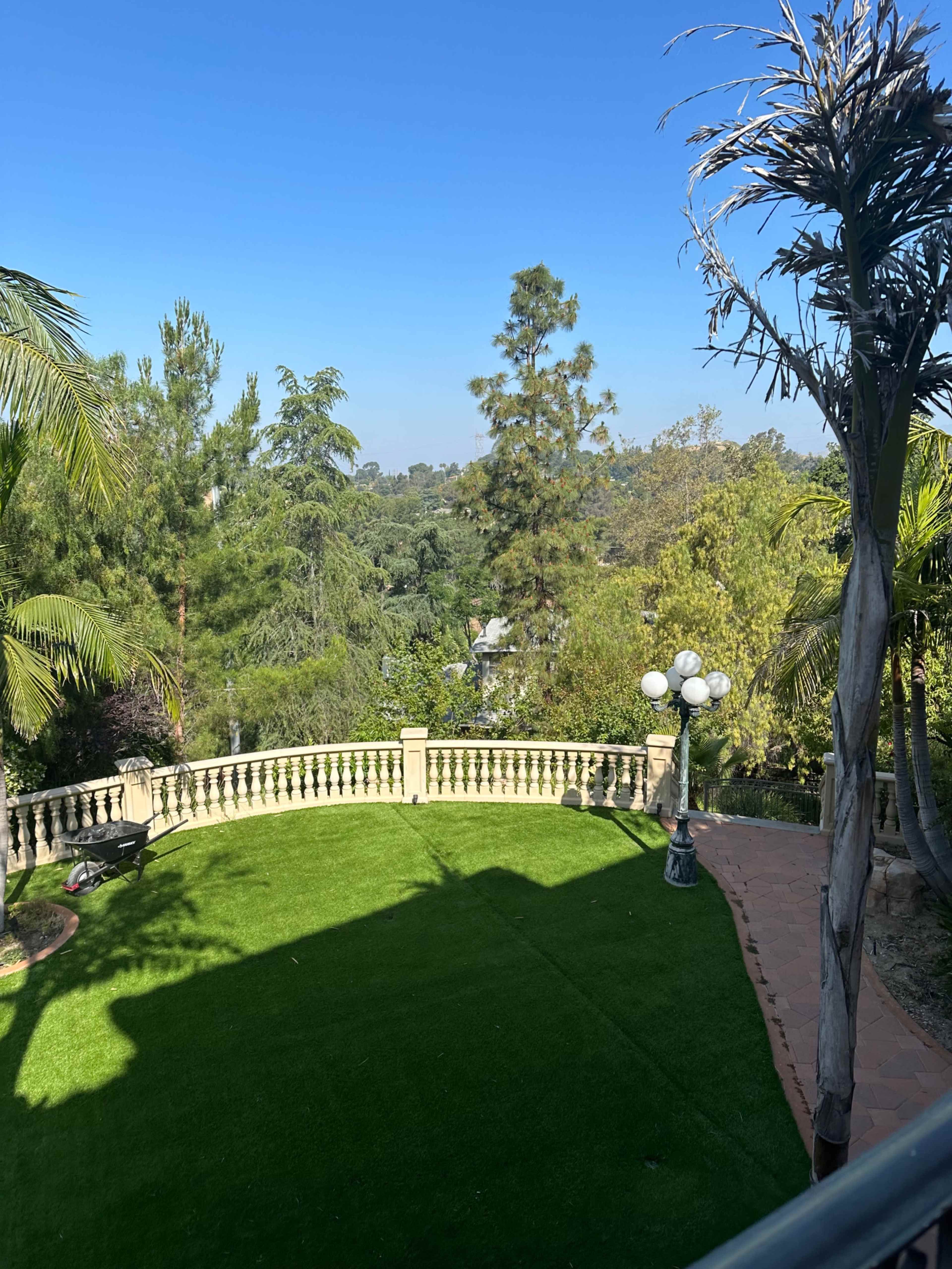 The image shows a well-maintained green lawn surrounded by lush trees and a decorative stone railing, with clear blue skies above.