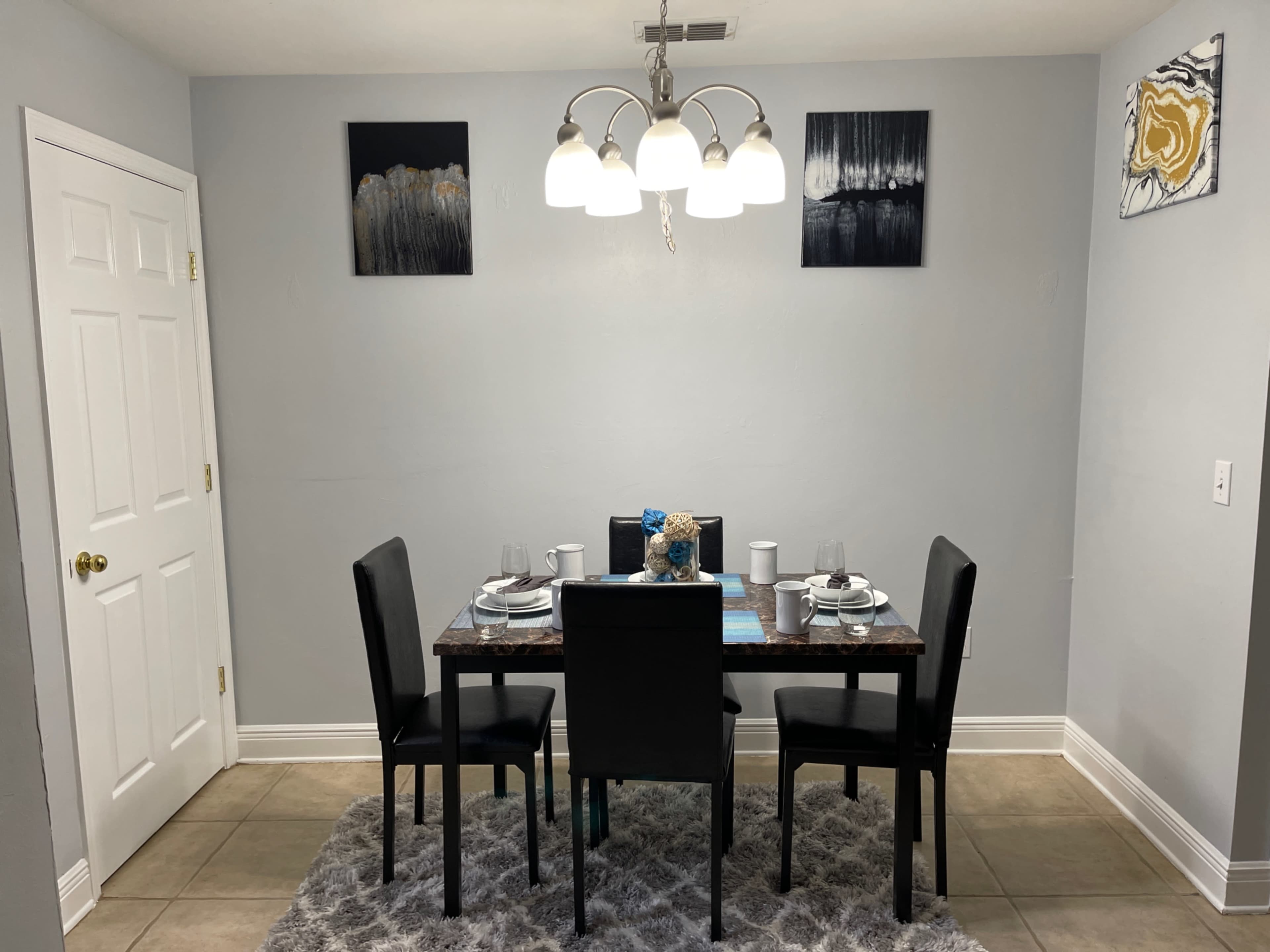 A dining area features a rectangular table set for four, surrounded by black chairs, with artwork on the gray walls and a light fixture above.