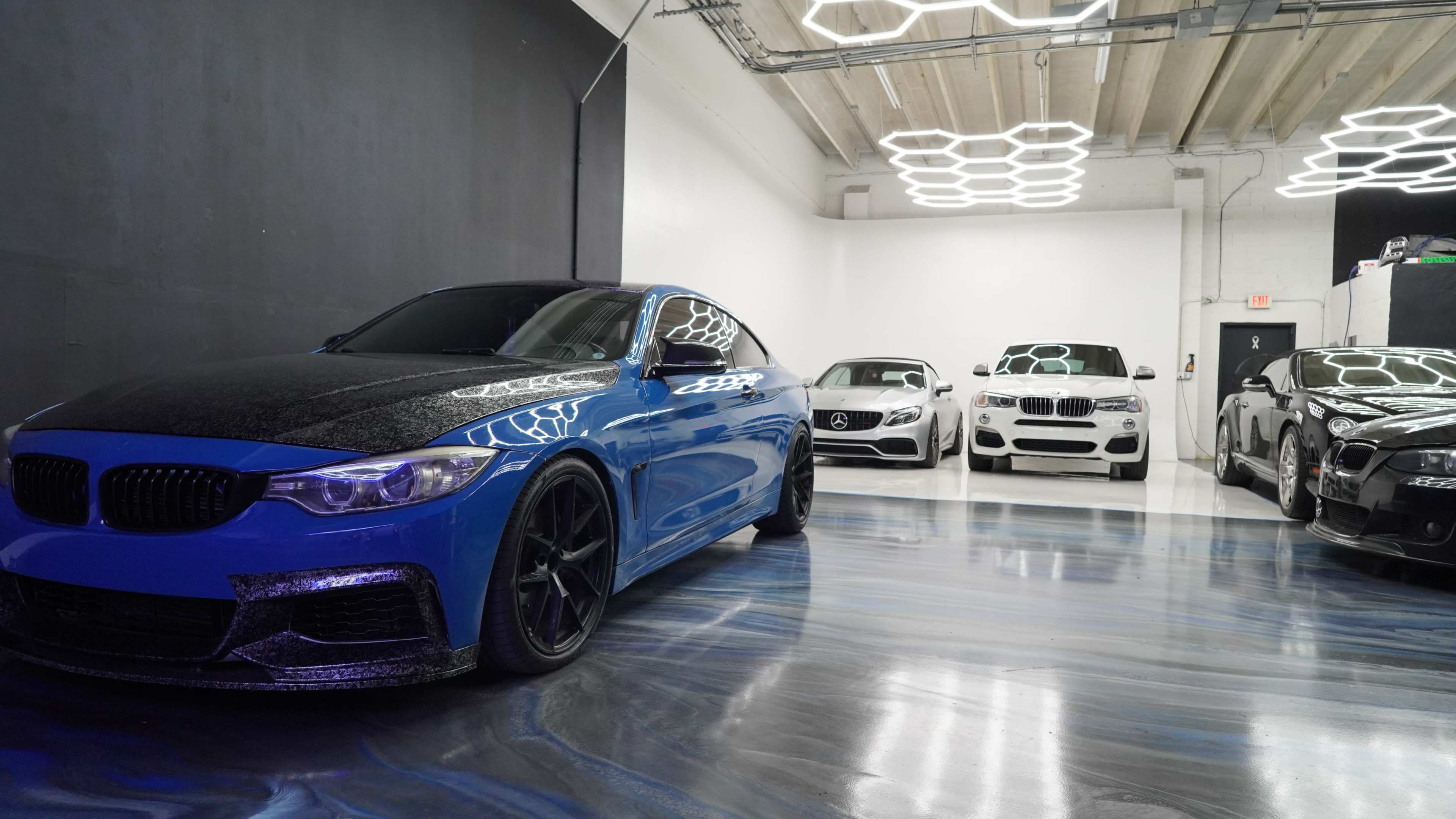 The image shows a sleek blue BMW with a carbon fiber hood parked in a modern car showroom alongside other luxury vehicles.