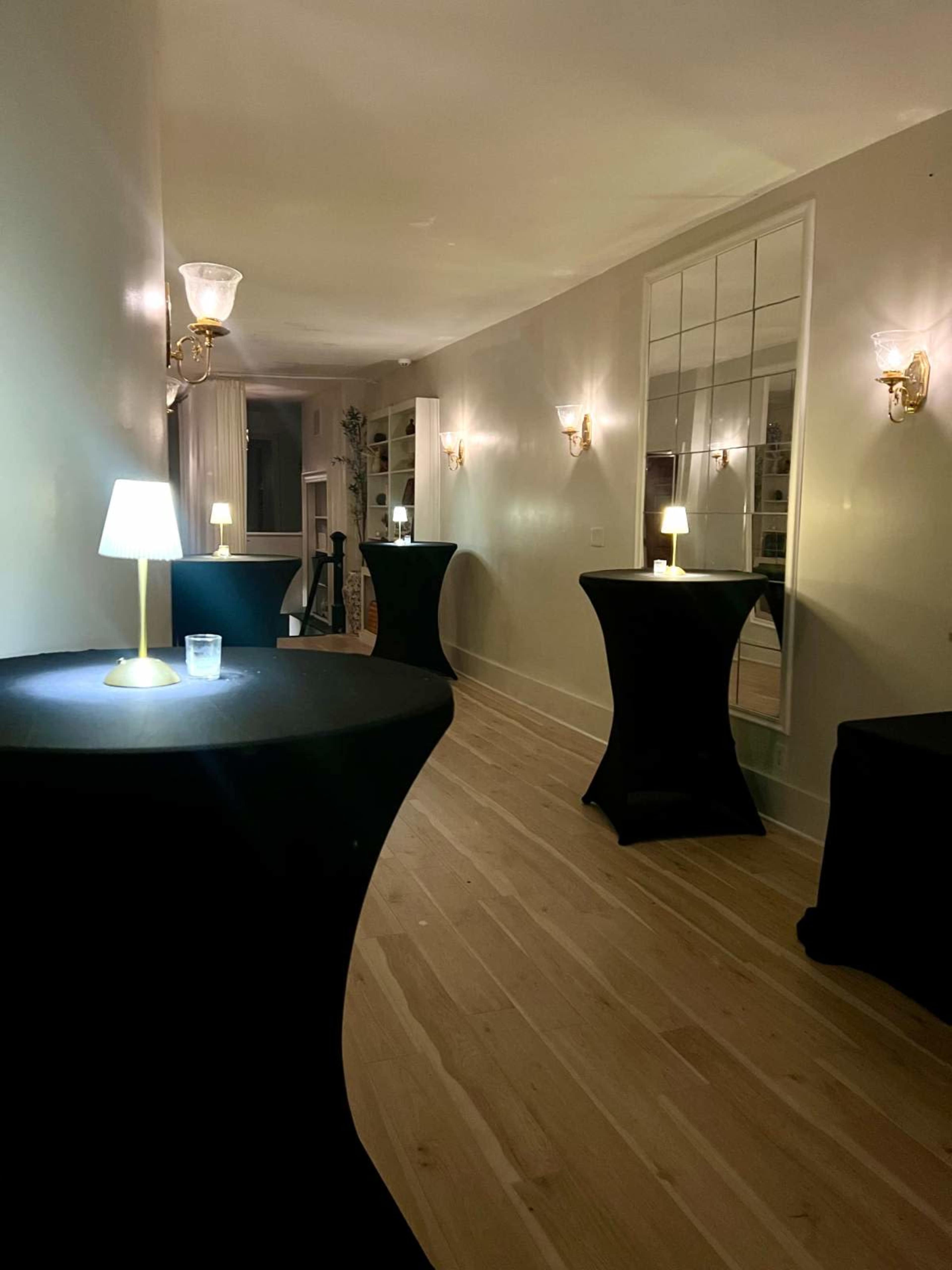 The image shows a dimly lit hallway with several tall, round tables covered in black cloth and small lamps on each table.