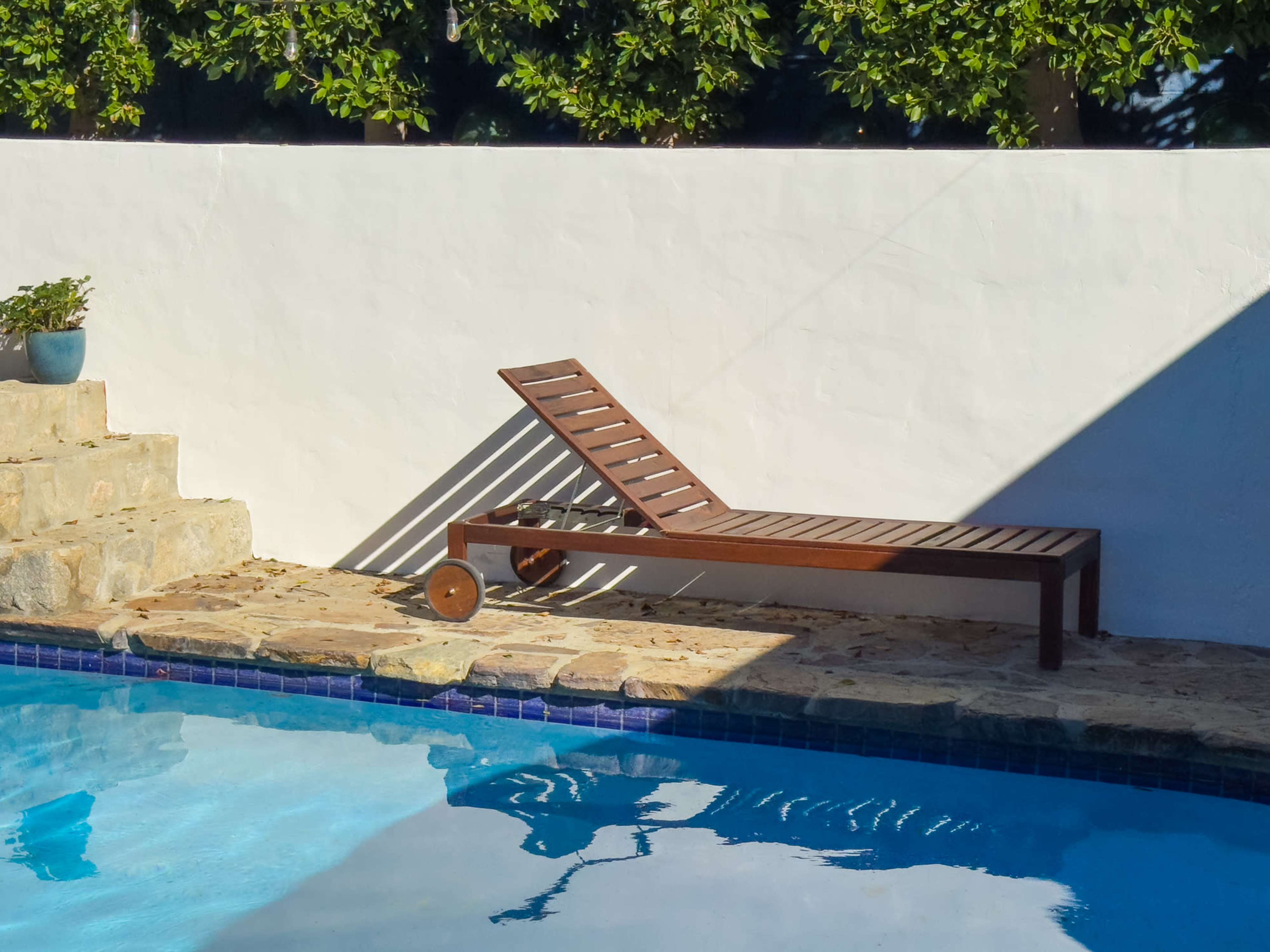 A wooden lounge chair is positioned beside a blue swimming pool with stone steps leading up to it, against a white wall.