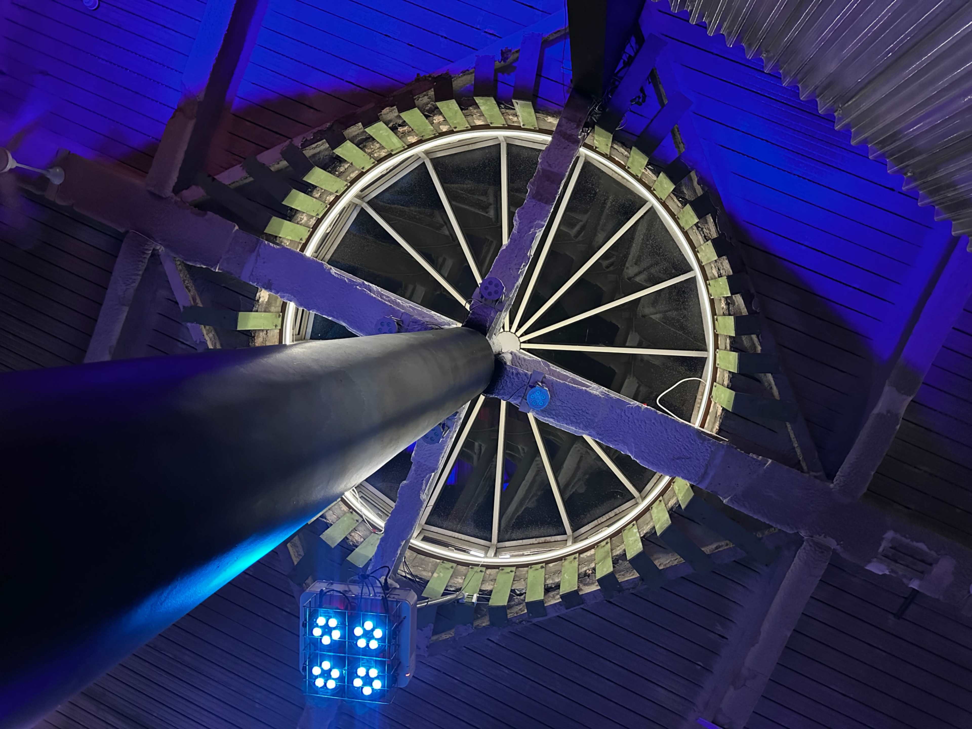 The image shows a circular structure viewed from below, featuring a central pole and a rim with evenly spaced green elements, illuminated by blue light.