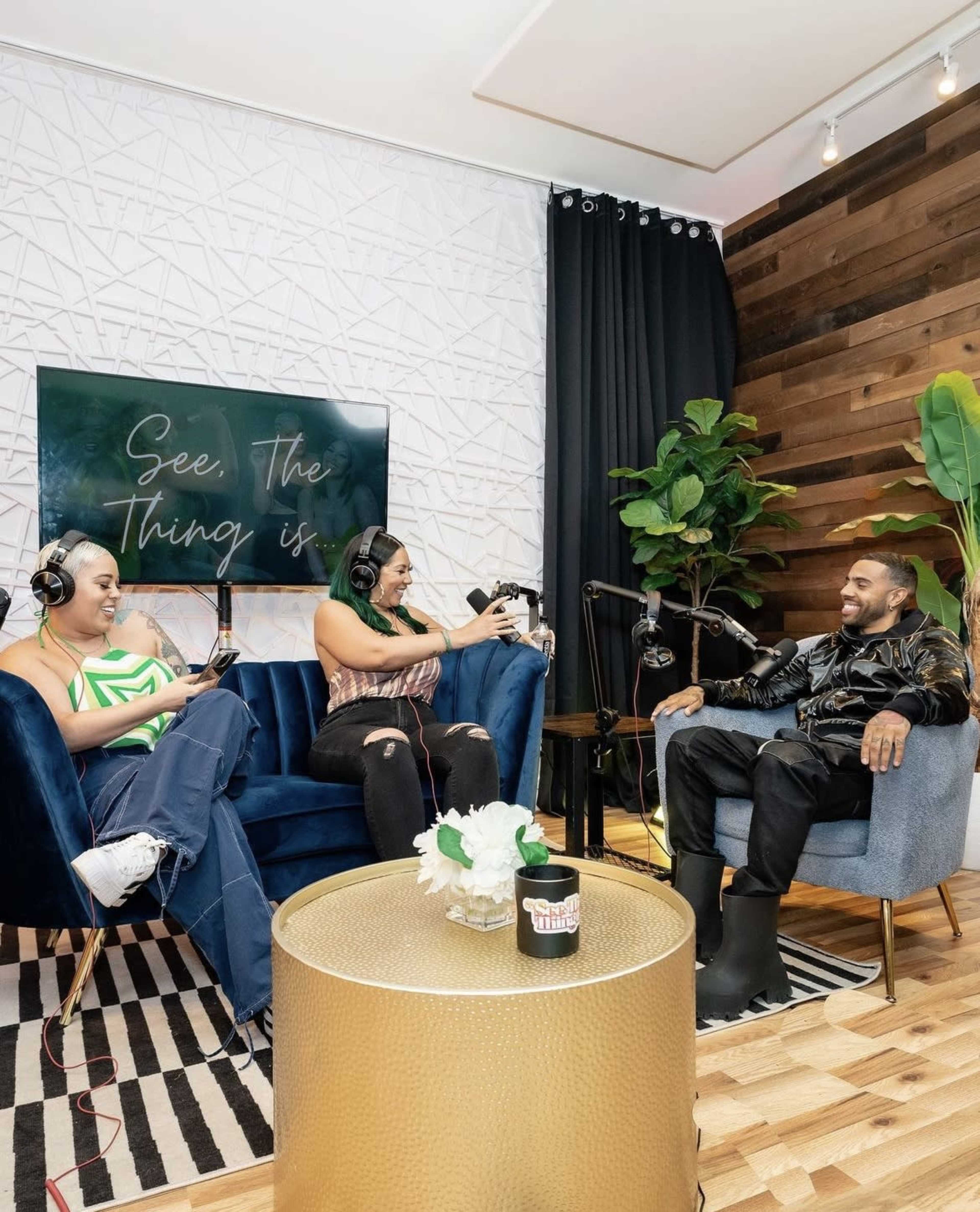 Two women and a man are seated in a well-decorated studio space, engaged in a conversation while using microphones.
