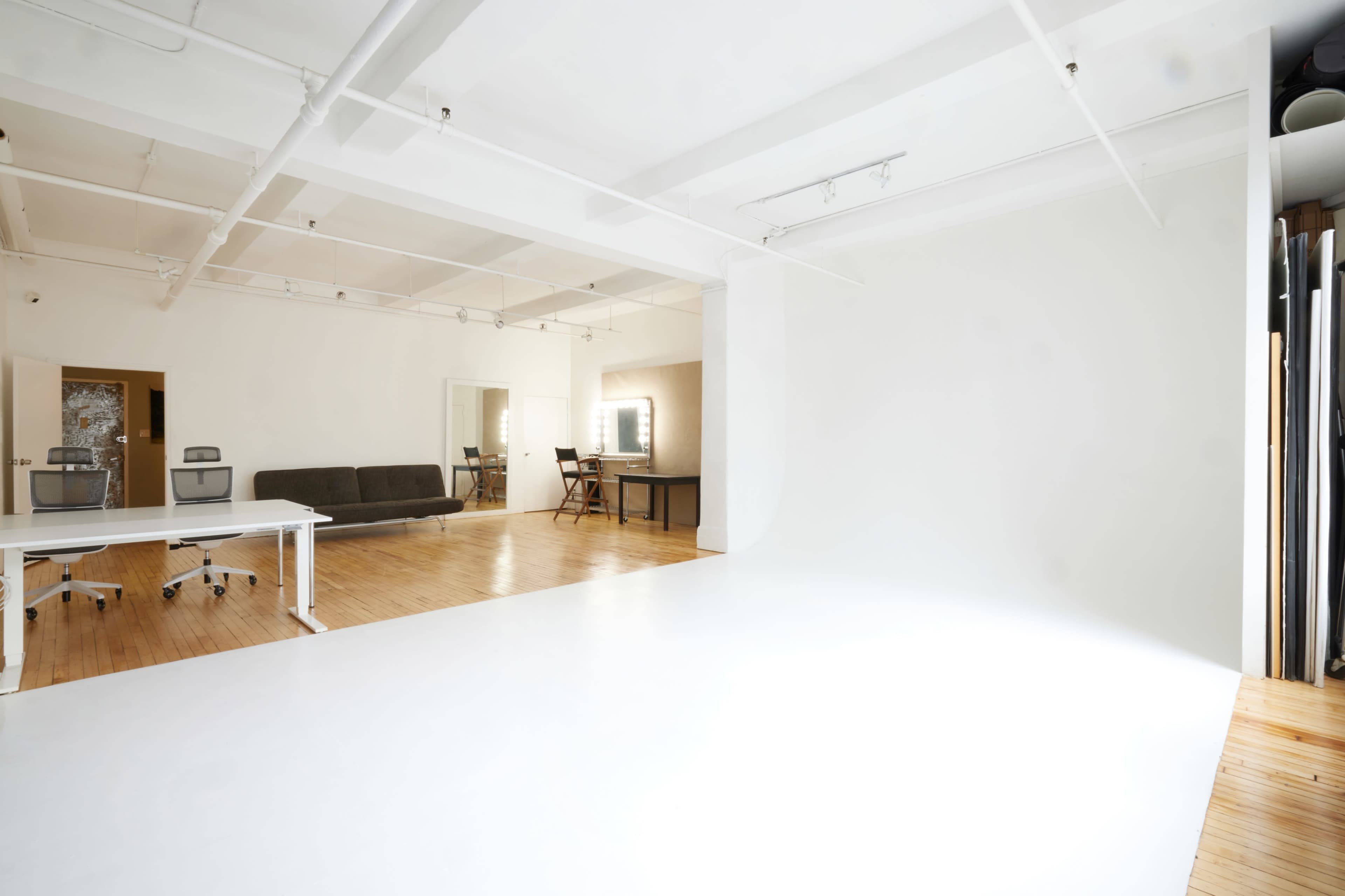 A spacious, minimalist studio with white walls, wooden flooring, and various furniture arrangements including a desk and a sofa.
