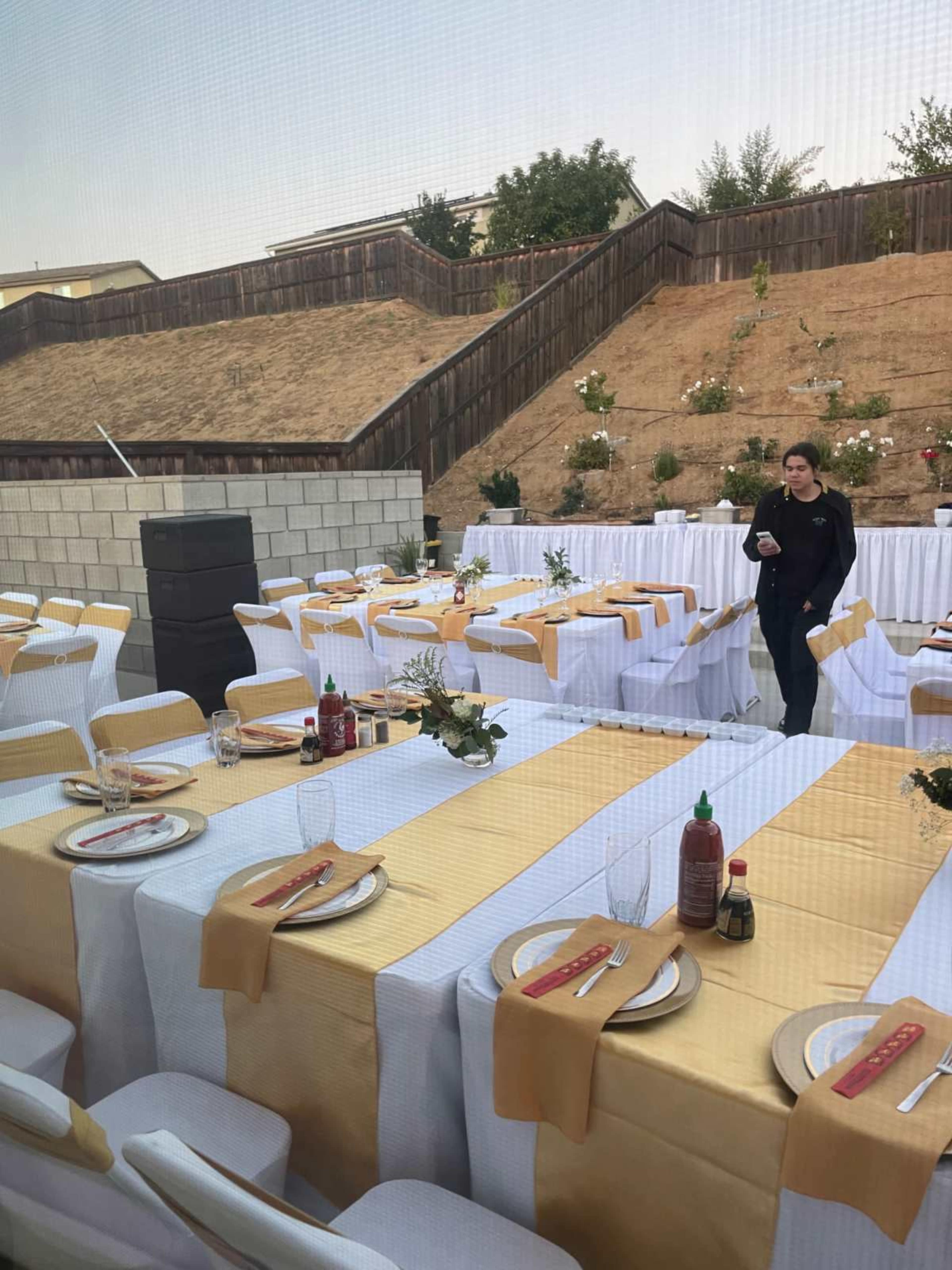 The image shows a banquet setup with several tables covered in white and yellow tablecloths, arranged outdoors against a backdrop of sloped landscaping.
