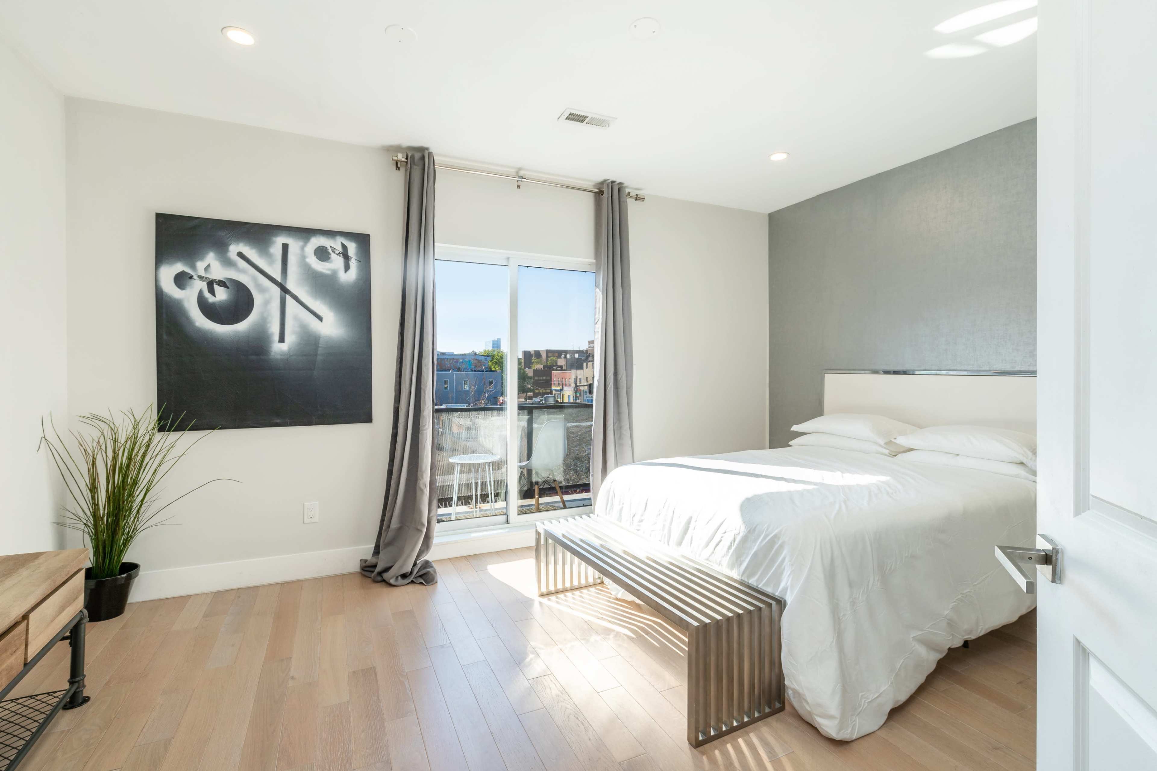A modern bedroom featuring a bed with white linens, a sliding glass door leading to a balcony, and a piece of abstract art on the wall.