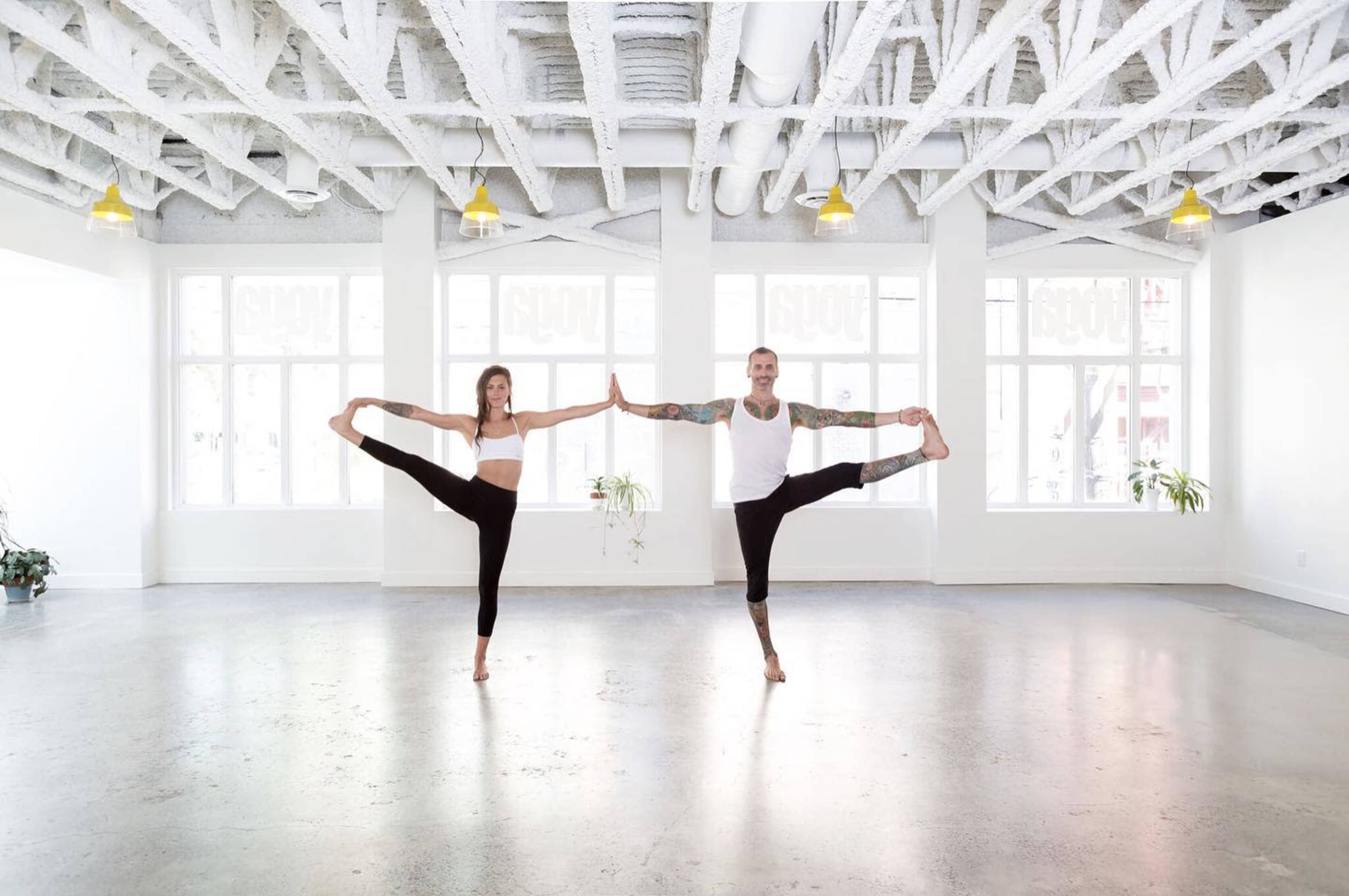 Two individuals are performing yoga poses in a bright, spacious studio with large windows and a minimalist design.