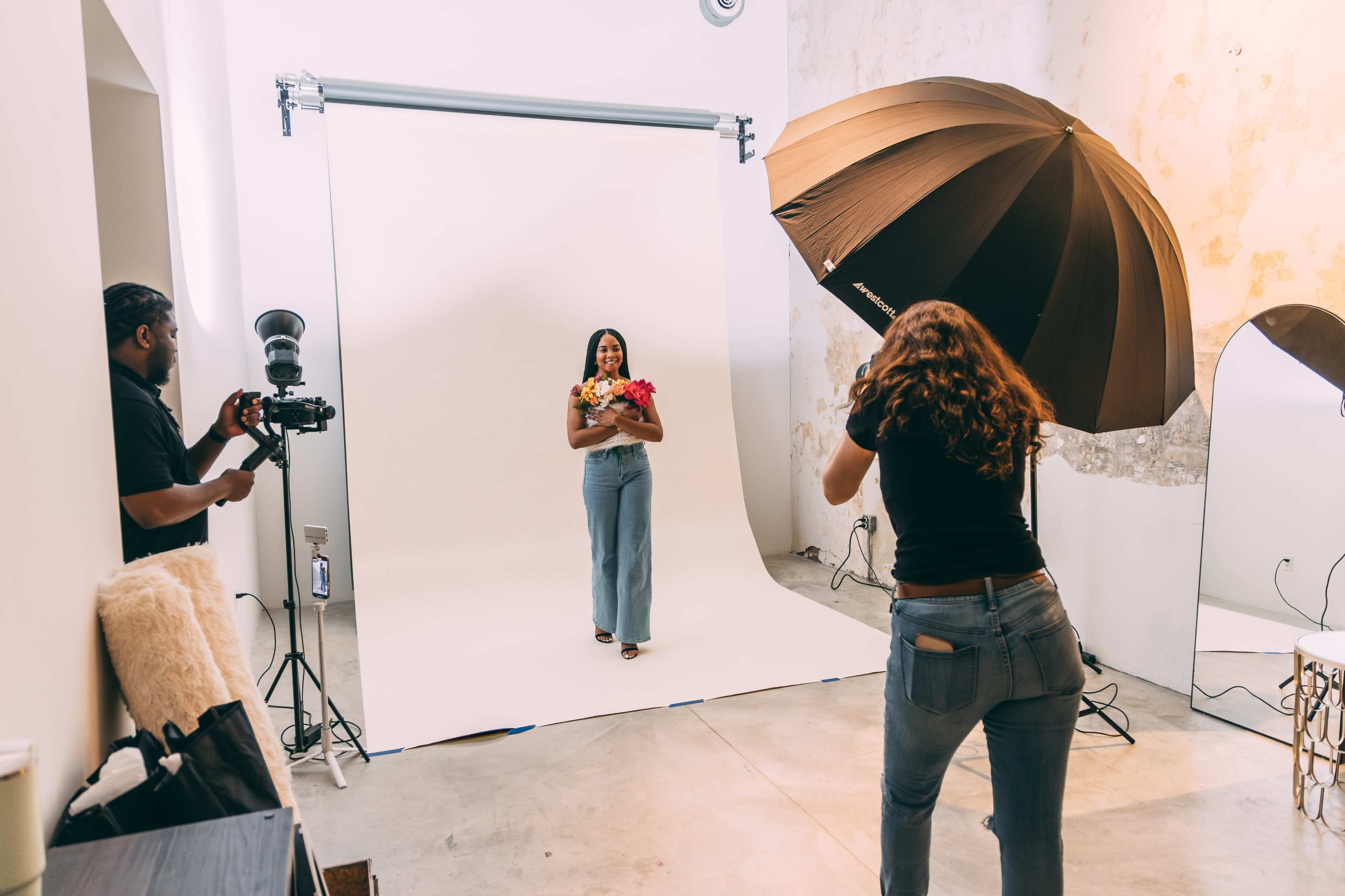 A woman stands holding flowers in front of a plain backdrop while a photographer and an assistant set up lighting in a studio.