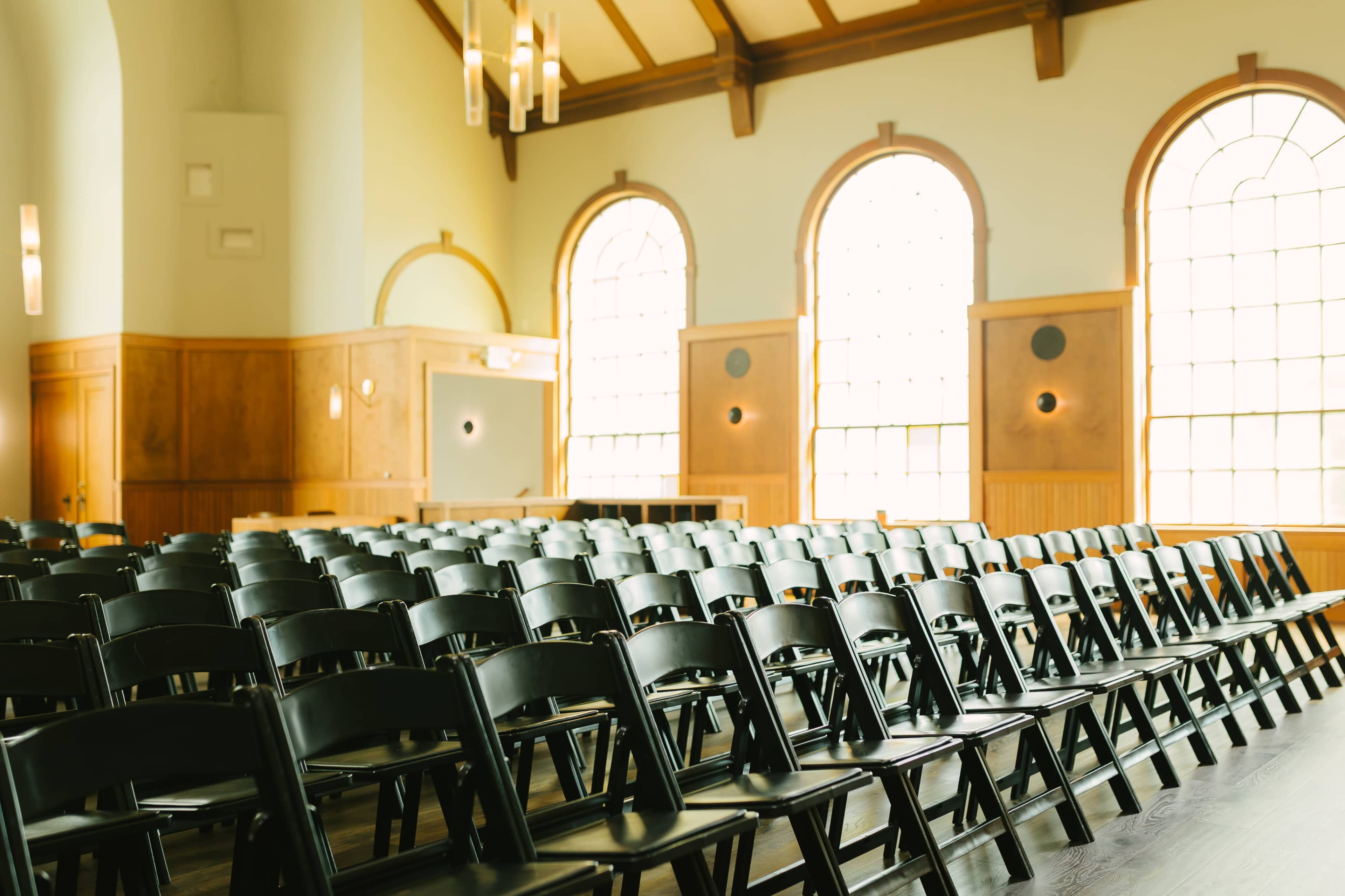 The image shows a room with rows of black folding chairs arranged neatly in front of large arched windows.