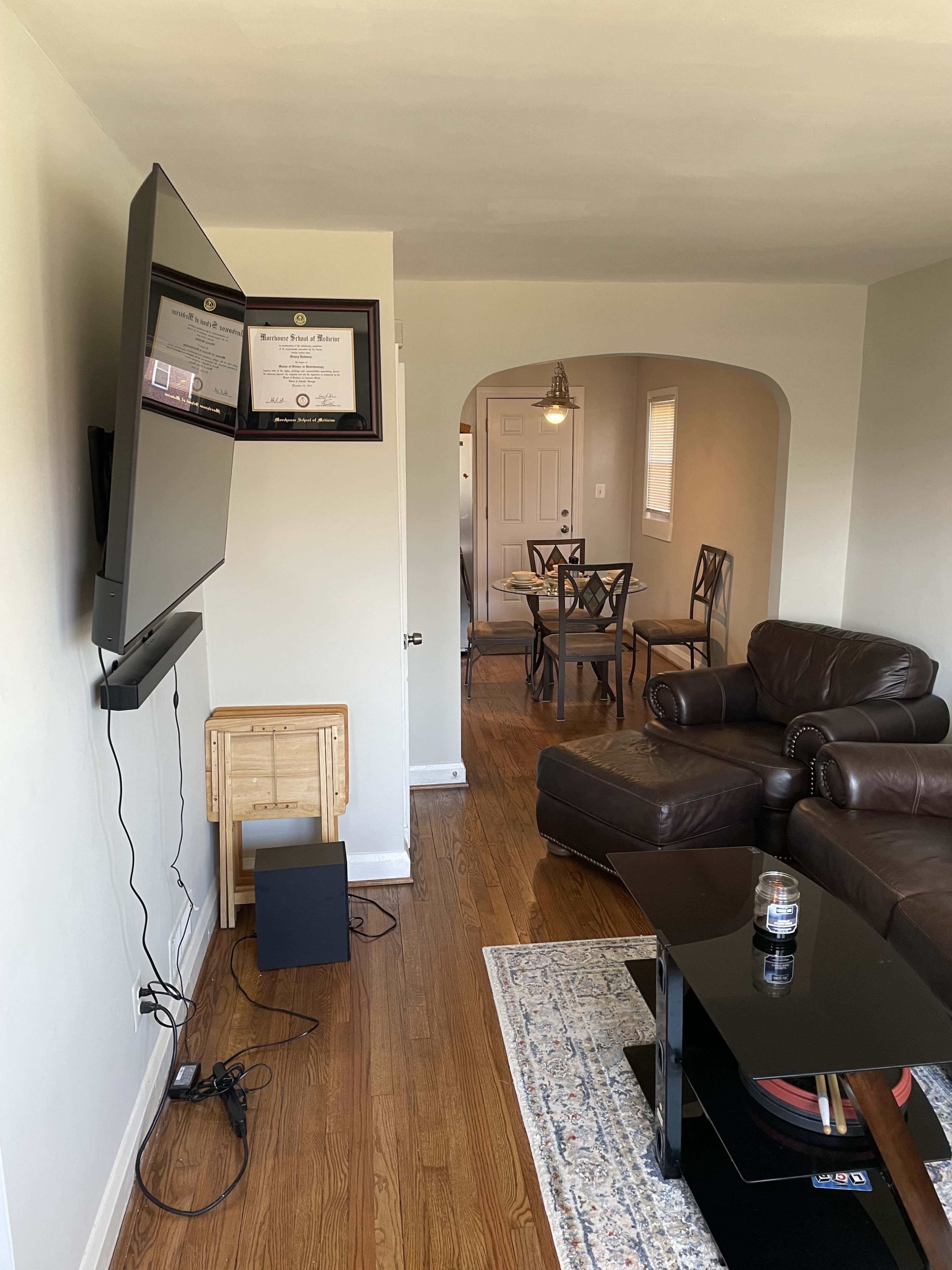 A living room features a wall-mounted television, a coffee table, a leather couch, and a dining area visible through an archway.