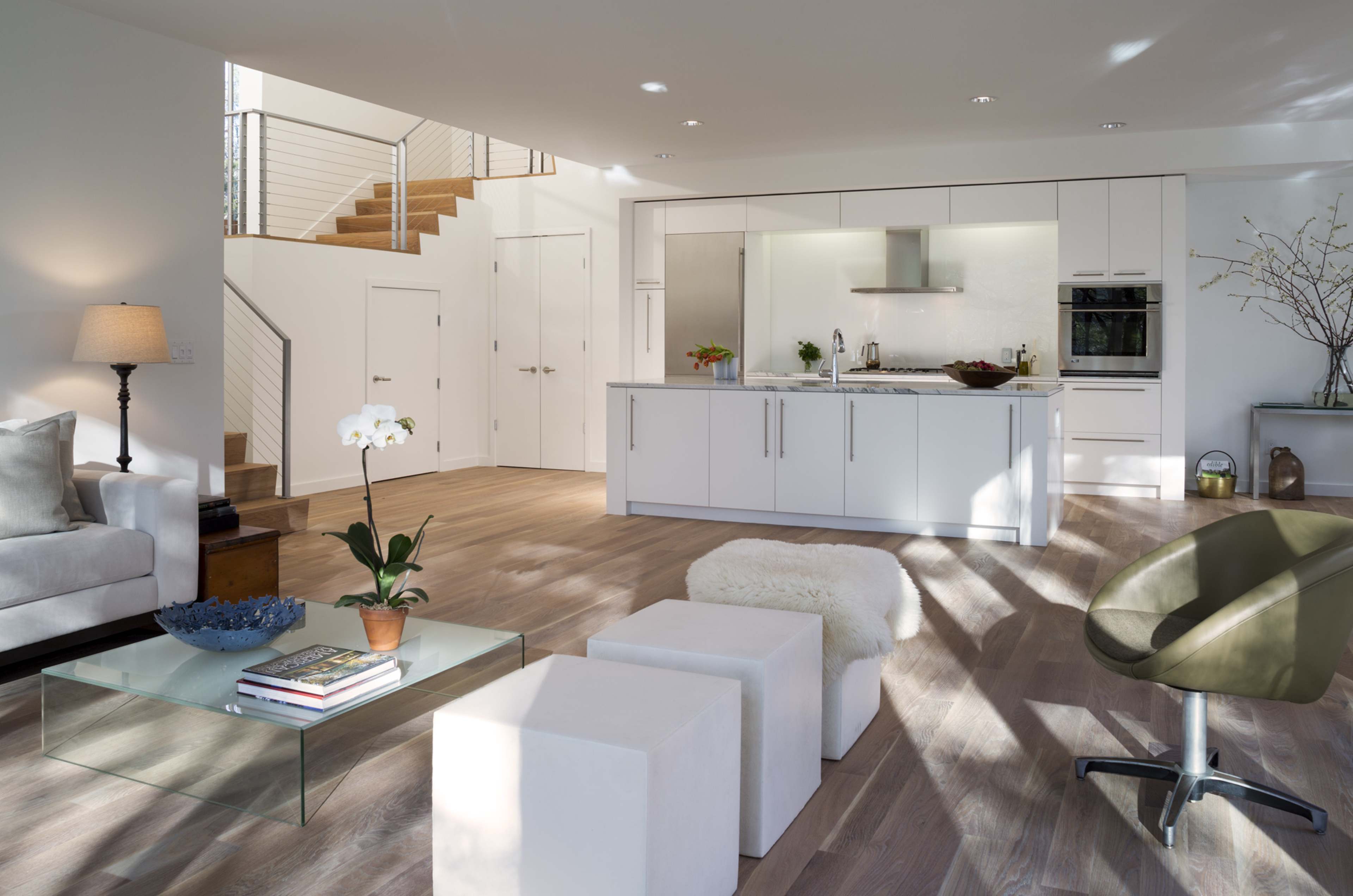 The image shows a modern, open-concept living space featuring a kitchen with white cabinetry, a sitting area with a gray sofa, and a staircase in the background.
