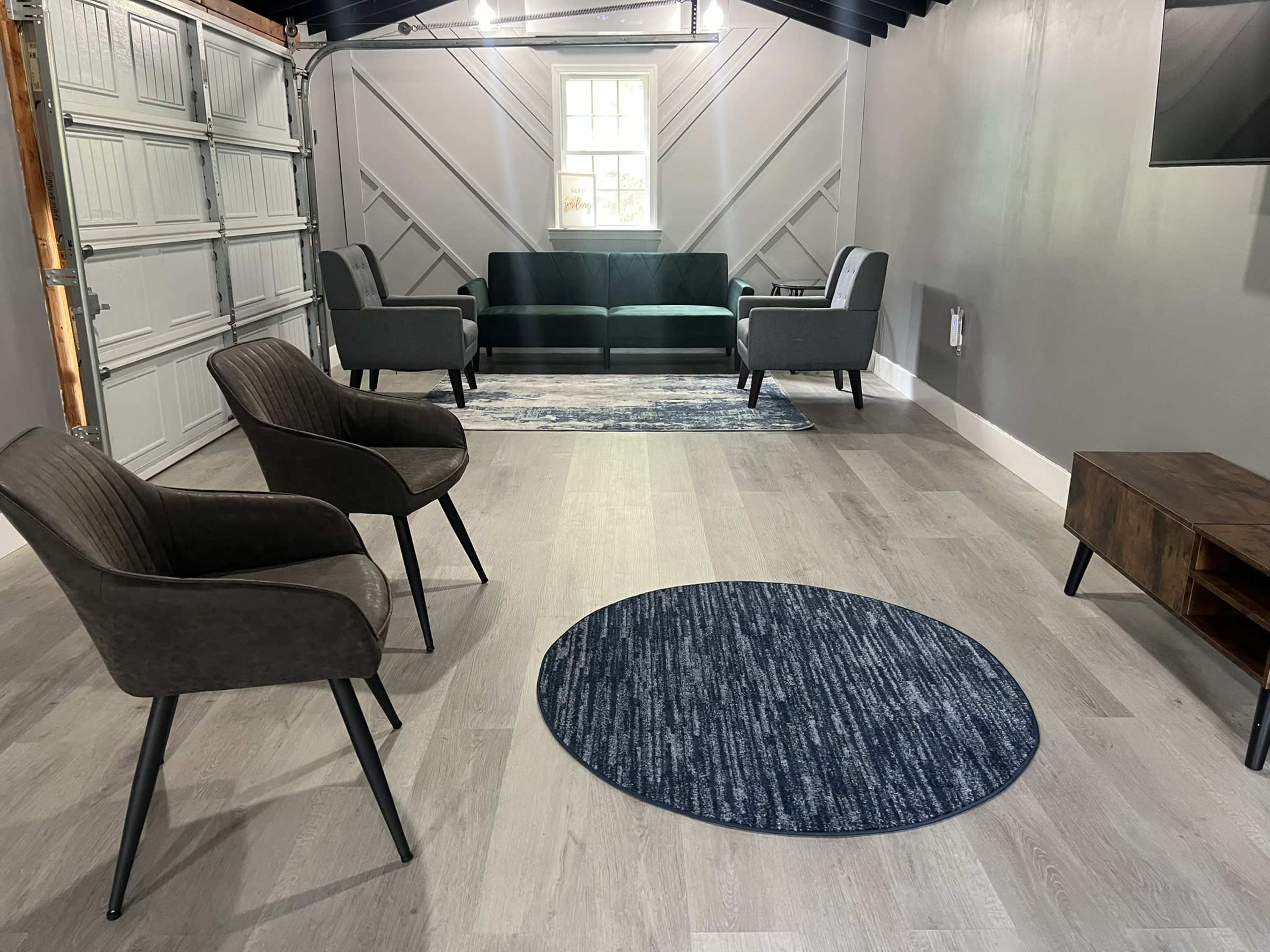 A modern, minimalist living space featuring a dark sofa, two armchairs, a circular rug, and a television mounted on the wall, all situated in a garage with a grey interior.