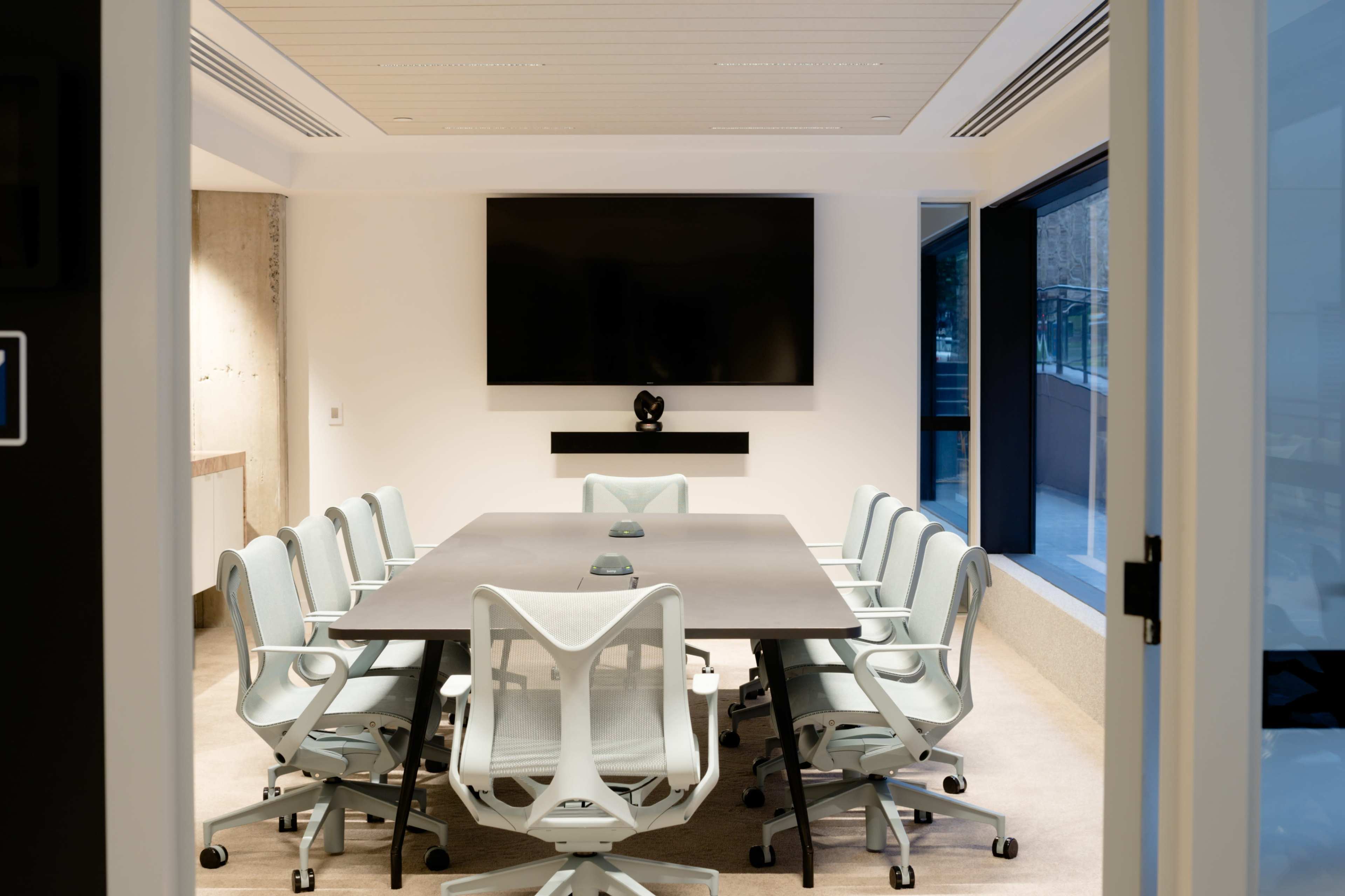 A modern conference room features a long table with twelve ergonomic chairs, a wall-mounted television, and large windows.
