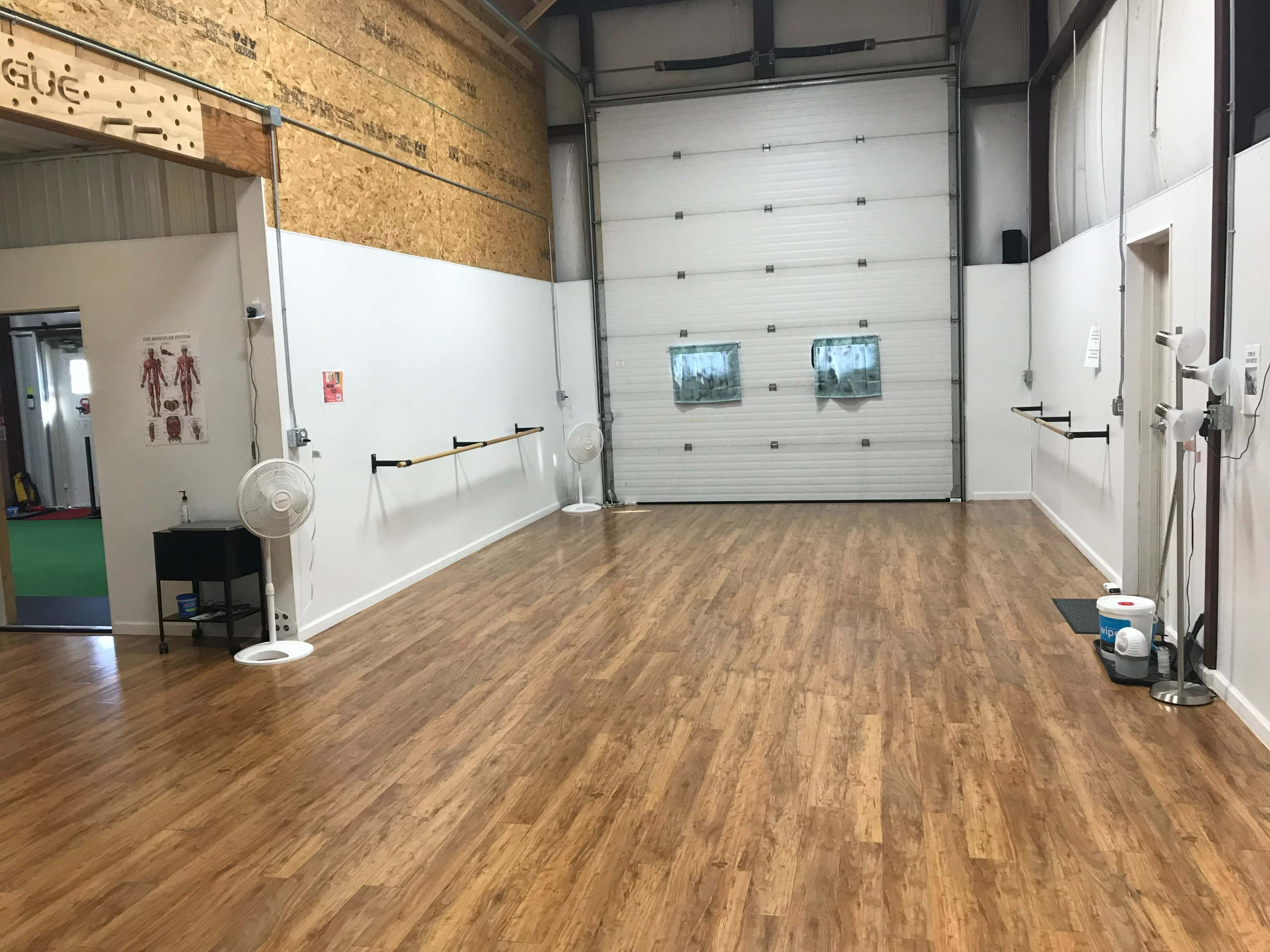 An interior space with wooden flooring, a large garage door, wall-mounted fans, and ballet barres along one wall.