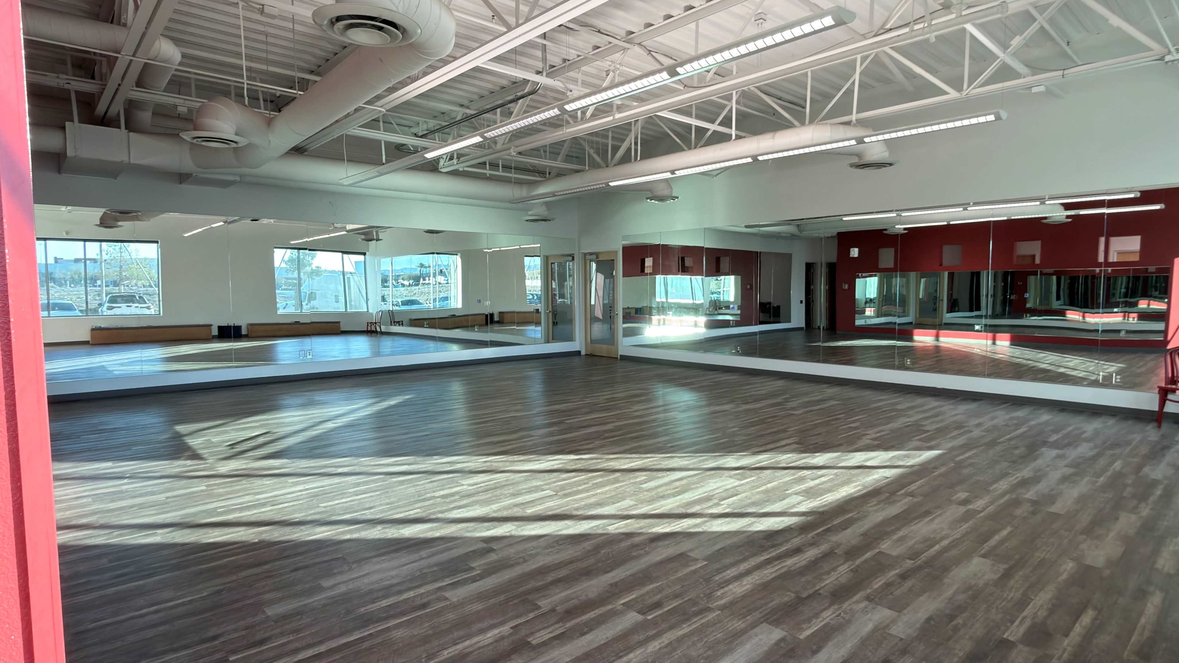 The image shows a spacious indoor studio with wooden flooring, large mirrors on the walls, and ample natural light coming through windows.