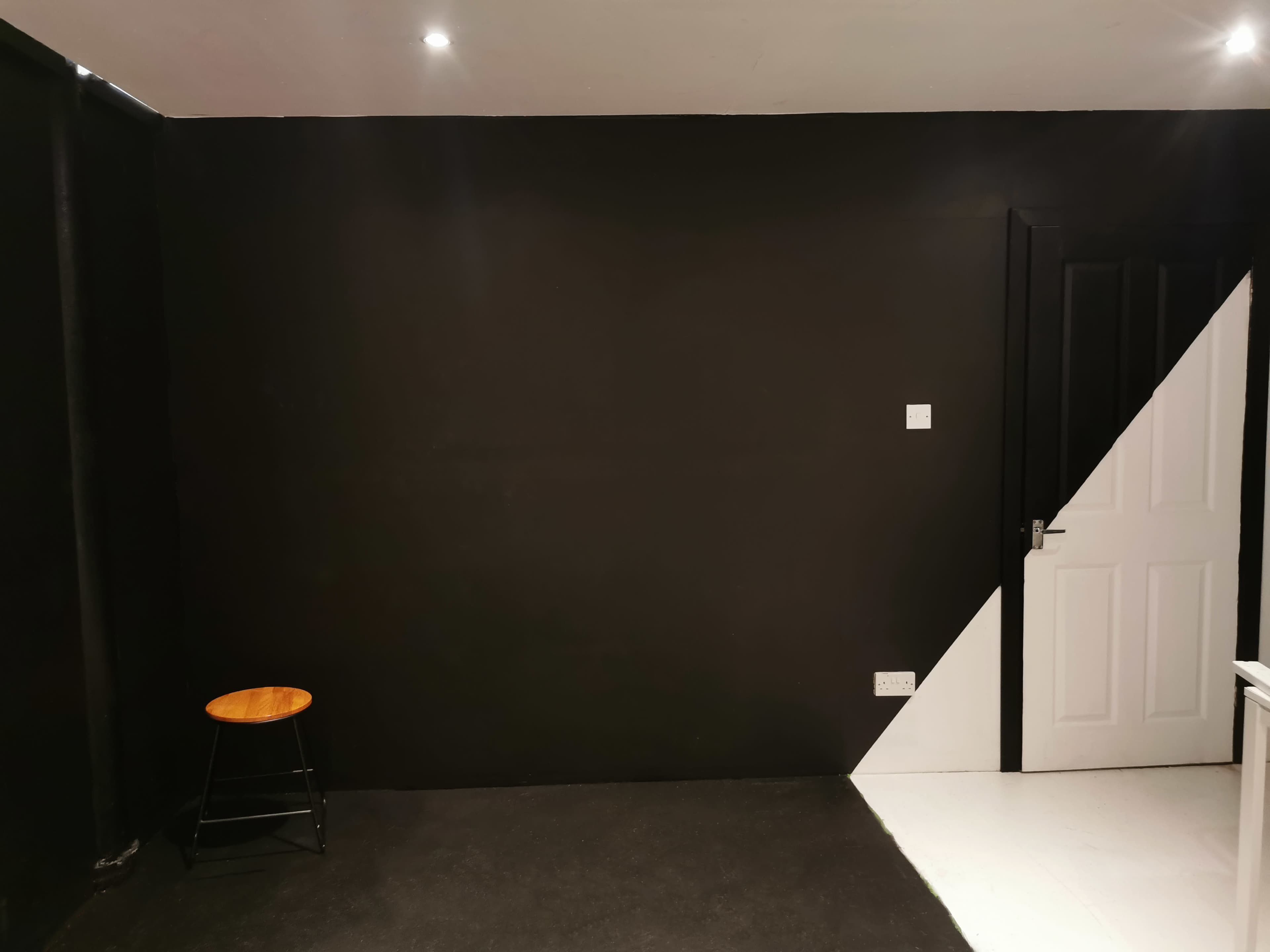 The image shows a room with two-tone painted walls, featuring a black wall on one side and a white wall on the other, along with a wooden stool and a closed door.