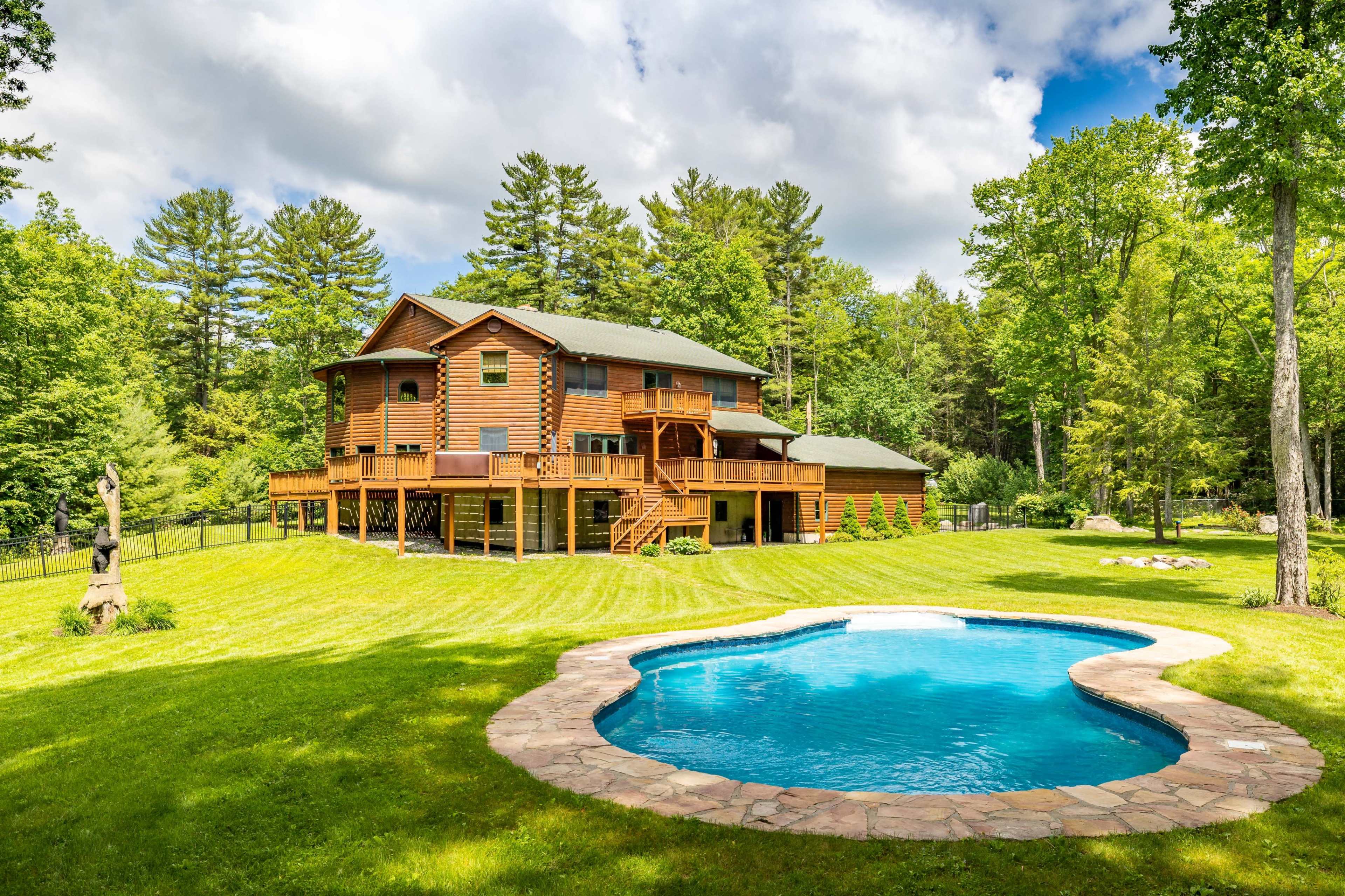 A log cabin-style house with multiple levels and a pool is surrounded by a lush green lawn and trees.