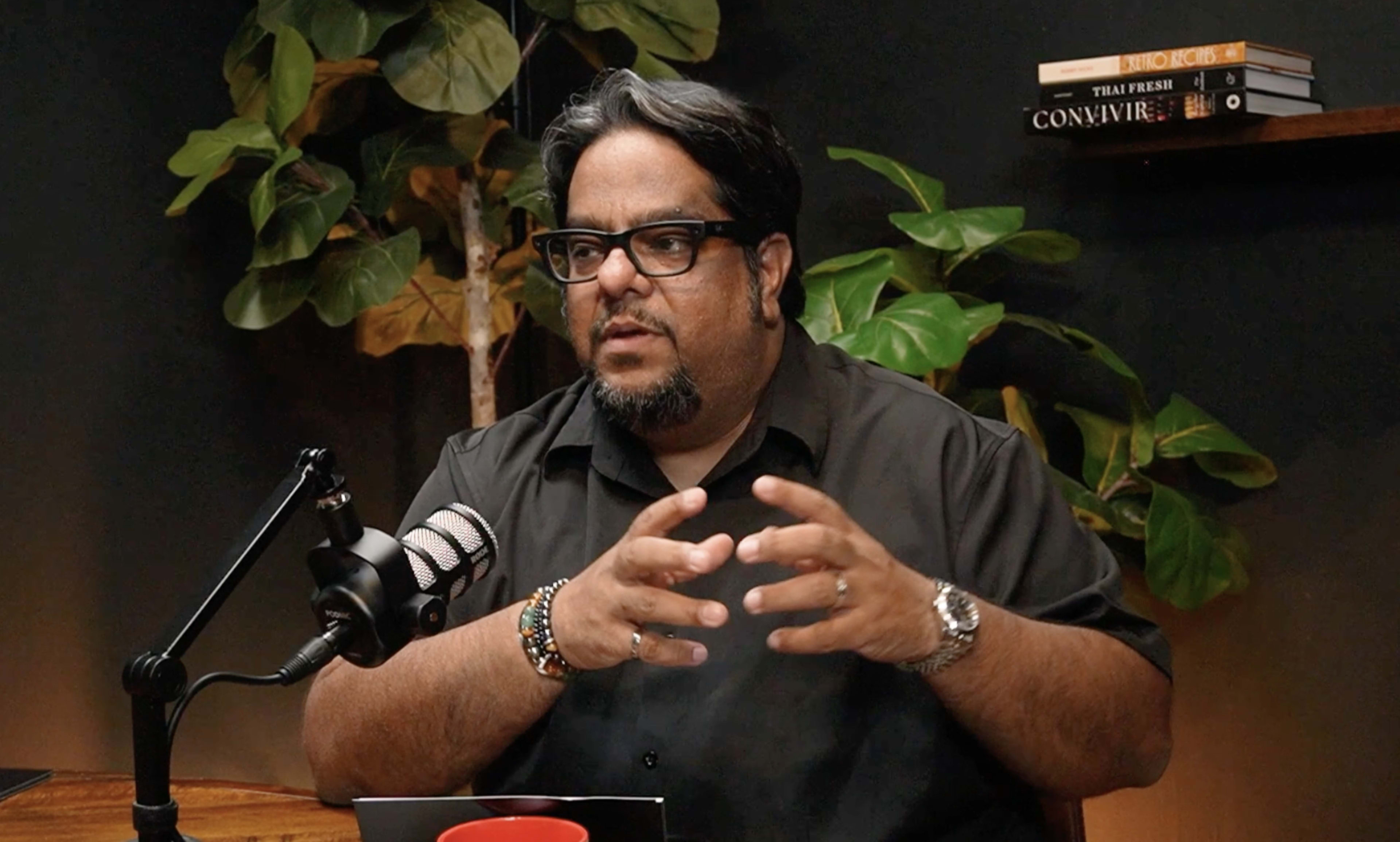 A person with long hair and glasses is speaking animatedly at a desk with a microphone, surrounded by potted plants and books on a shelf.