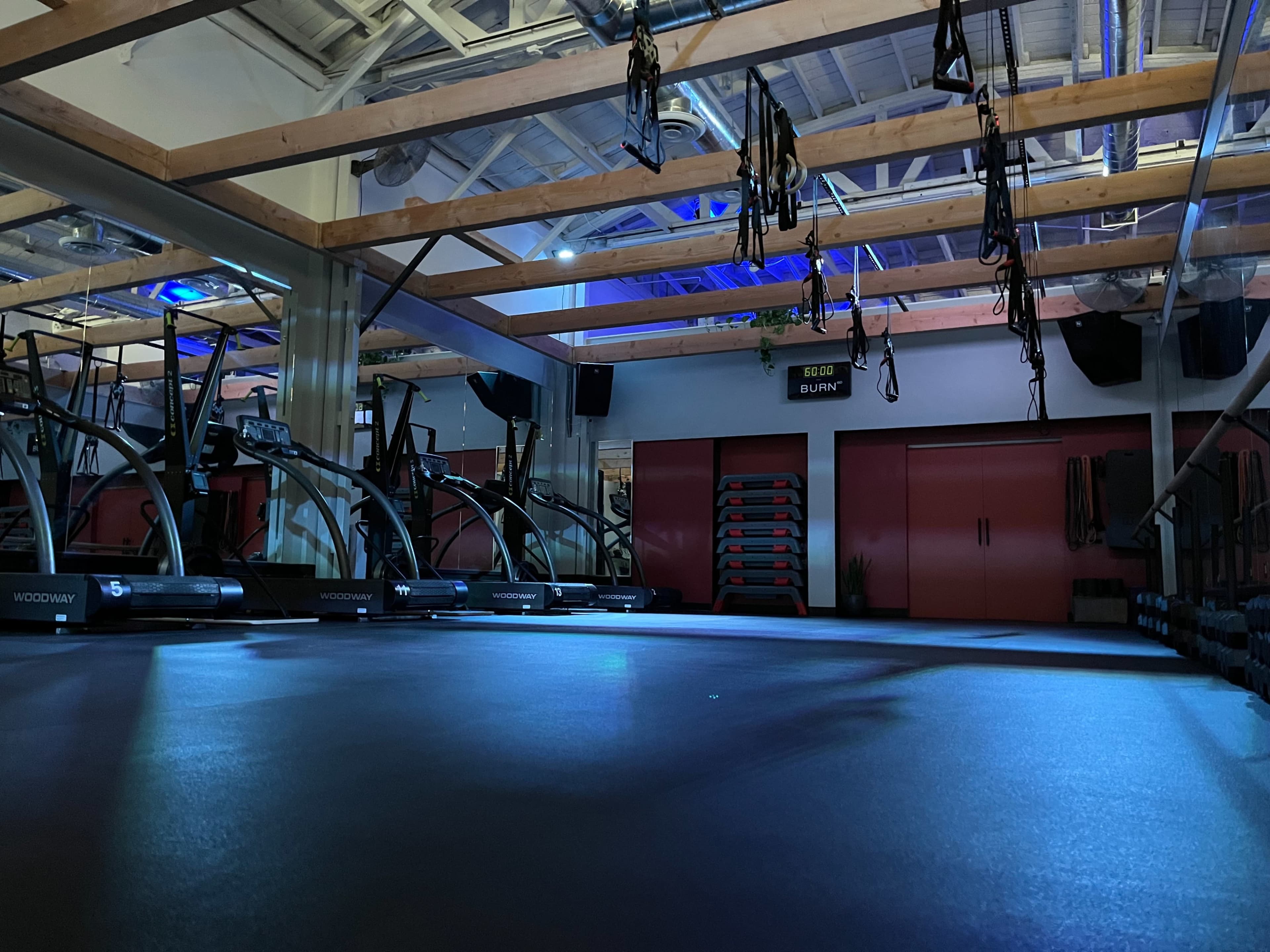 The image shows a dimly lit gym space equipped with exercise machines and suspended training ropes.