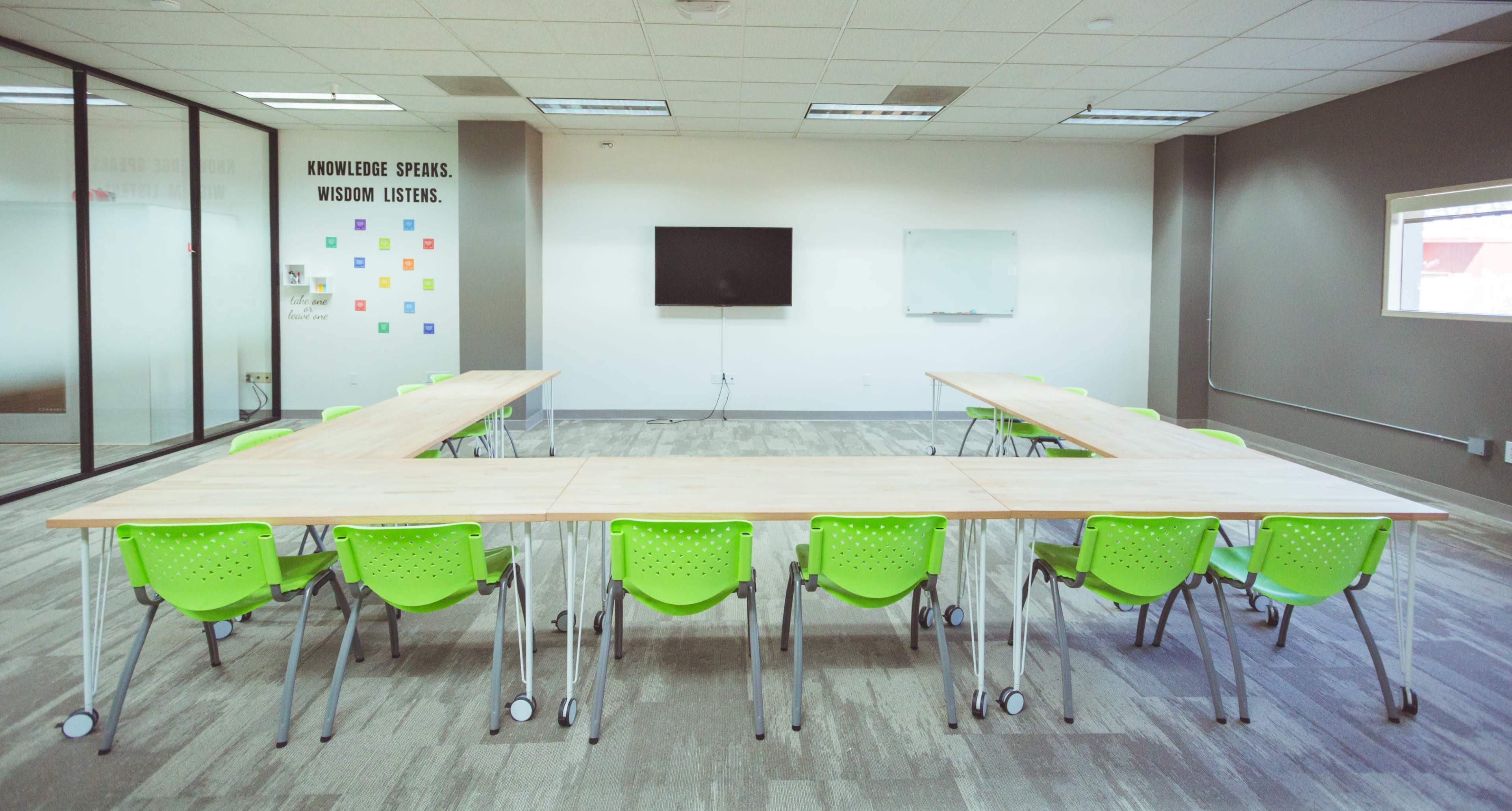 A modern conference room features a large, rectangular wooden table surrounded by green chairs, with a television and whiteboard on the walls.