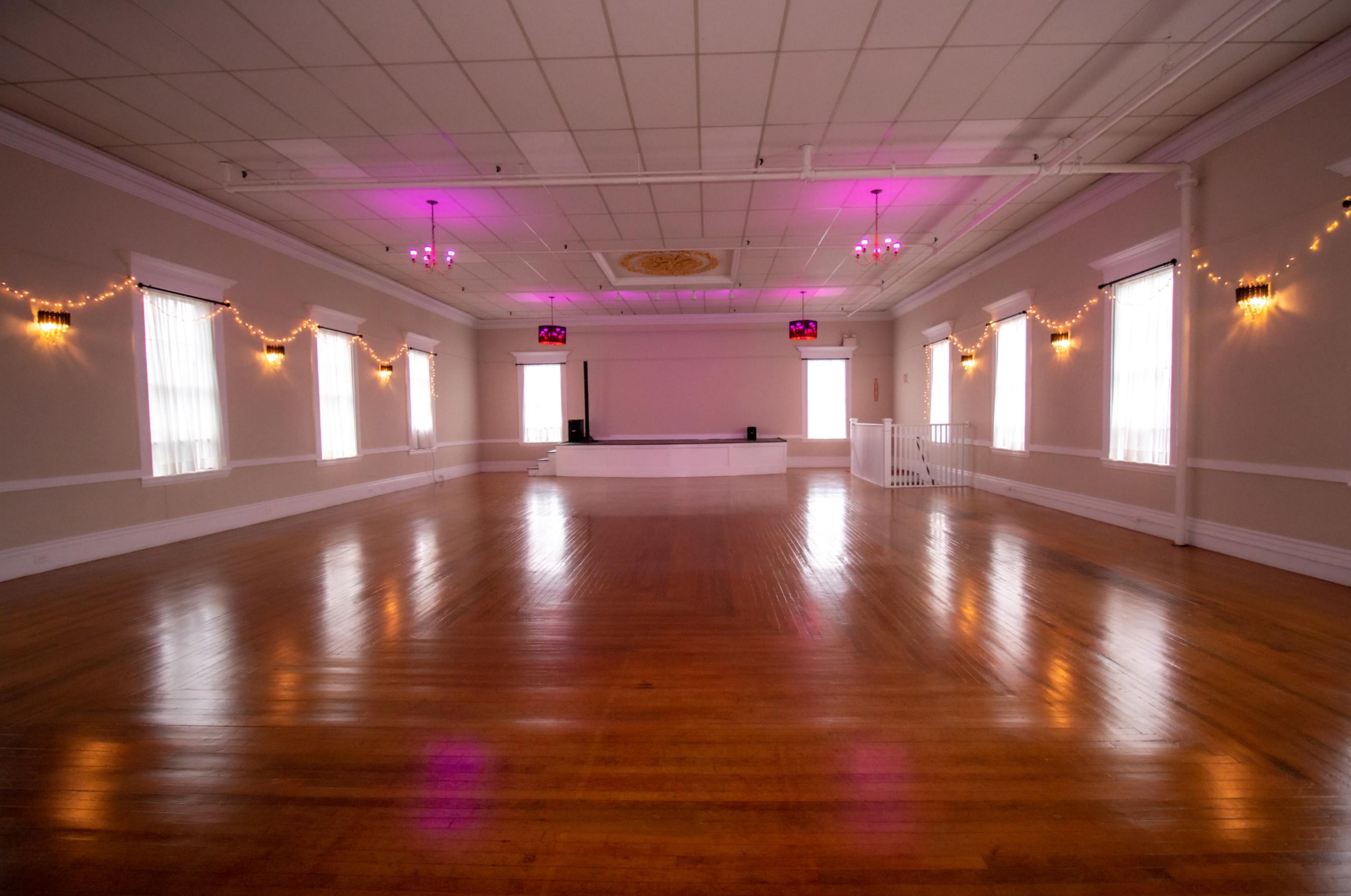 The image shows a spacious, well-lit event hall with a polished wooden floor, accented by pink lighting and decorative string lights along the walls.