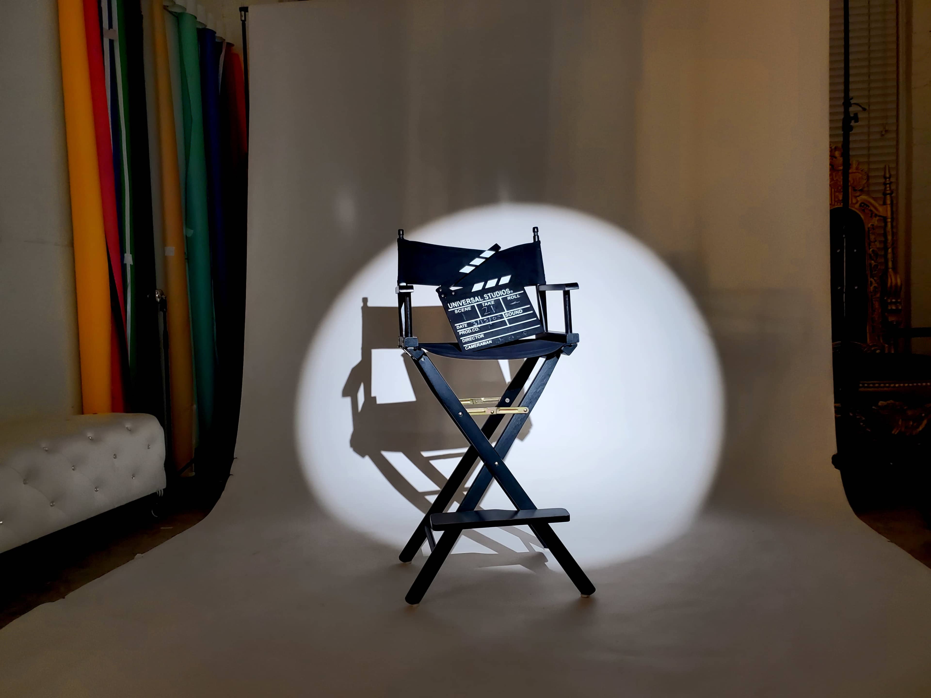 A director's chair is illuminated by a spotlight on a white backdrop, with colorful drapes positioned to the side.