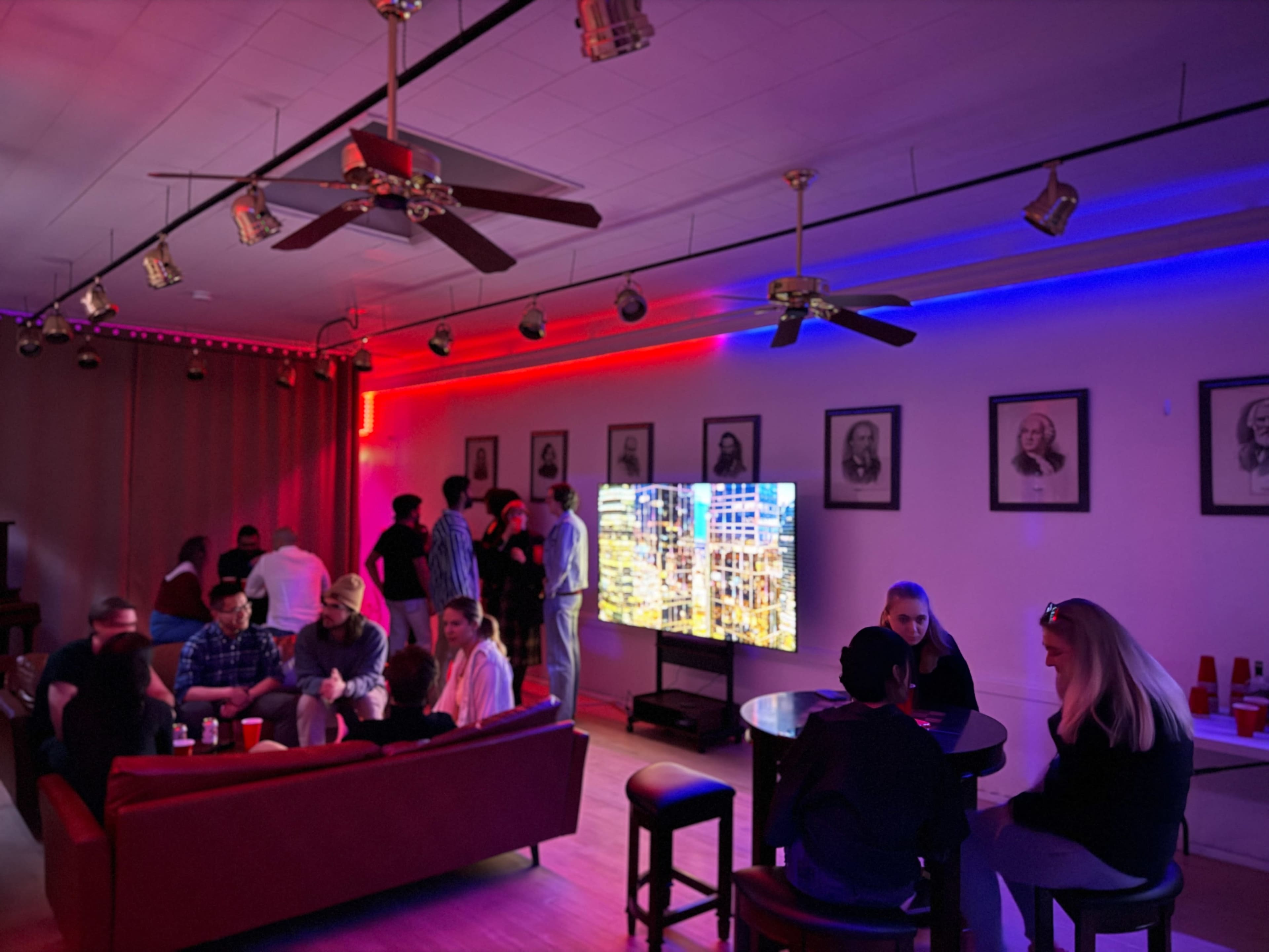 A group of people socializes in a well-lit room with blue and red accent lighting, featuring a large screen displaying images and framed portraits on the walls.
