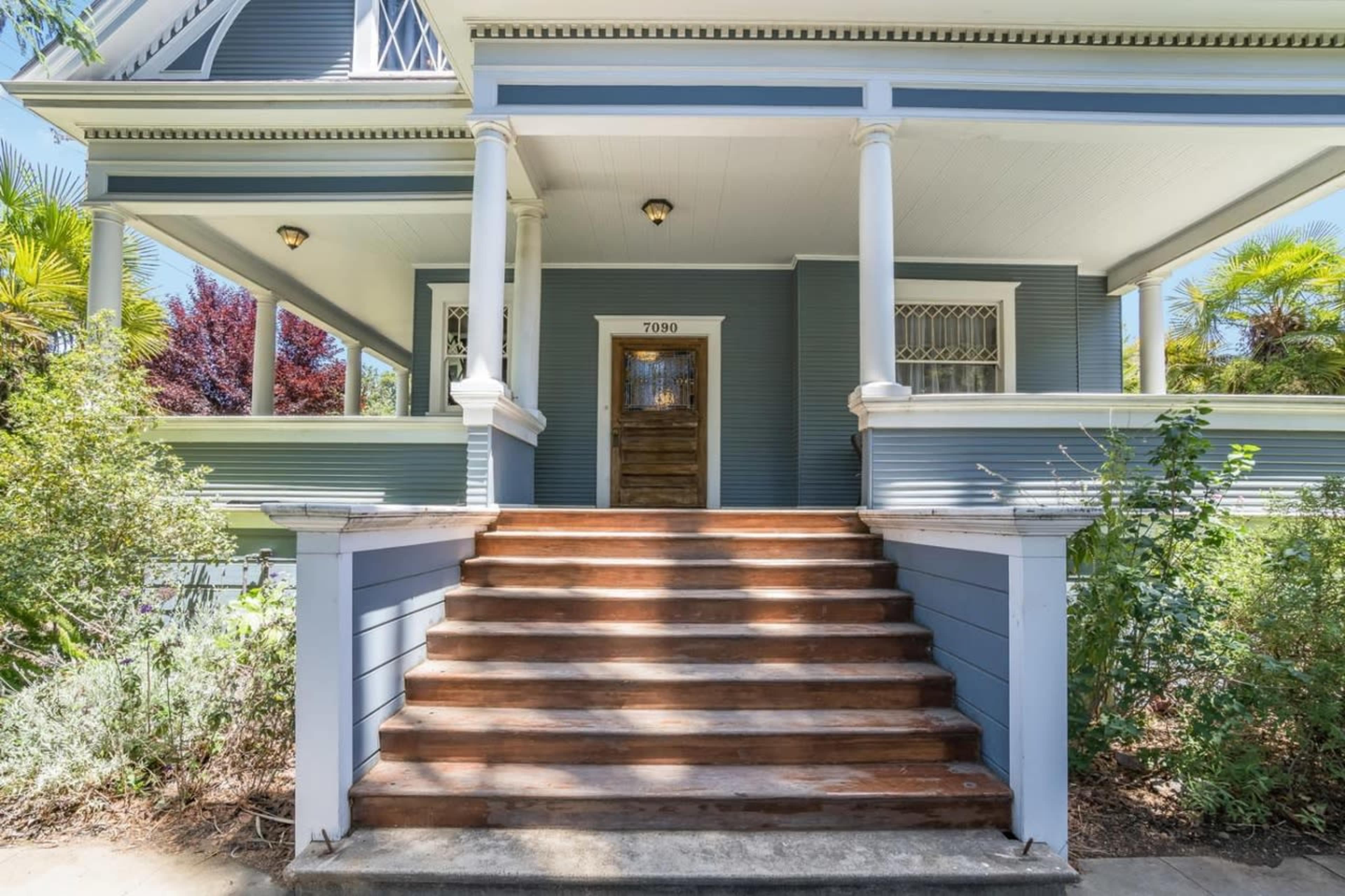 A set of wooden steps leads up to a blue house with a front door framed by white columns and greenery on either side.