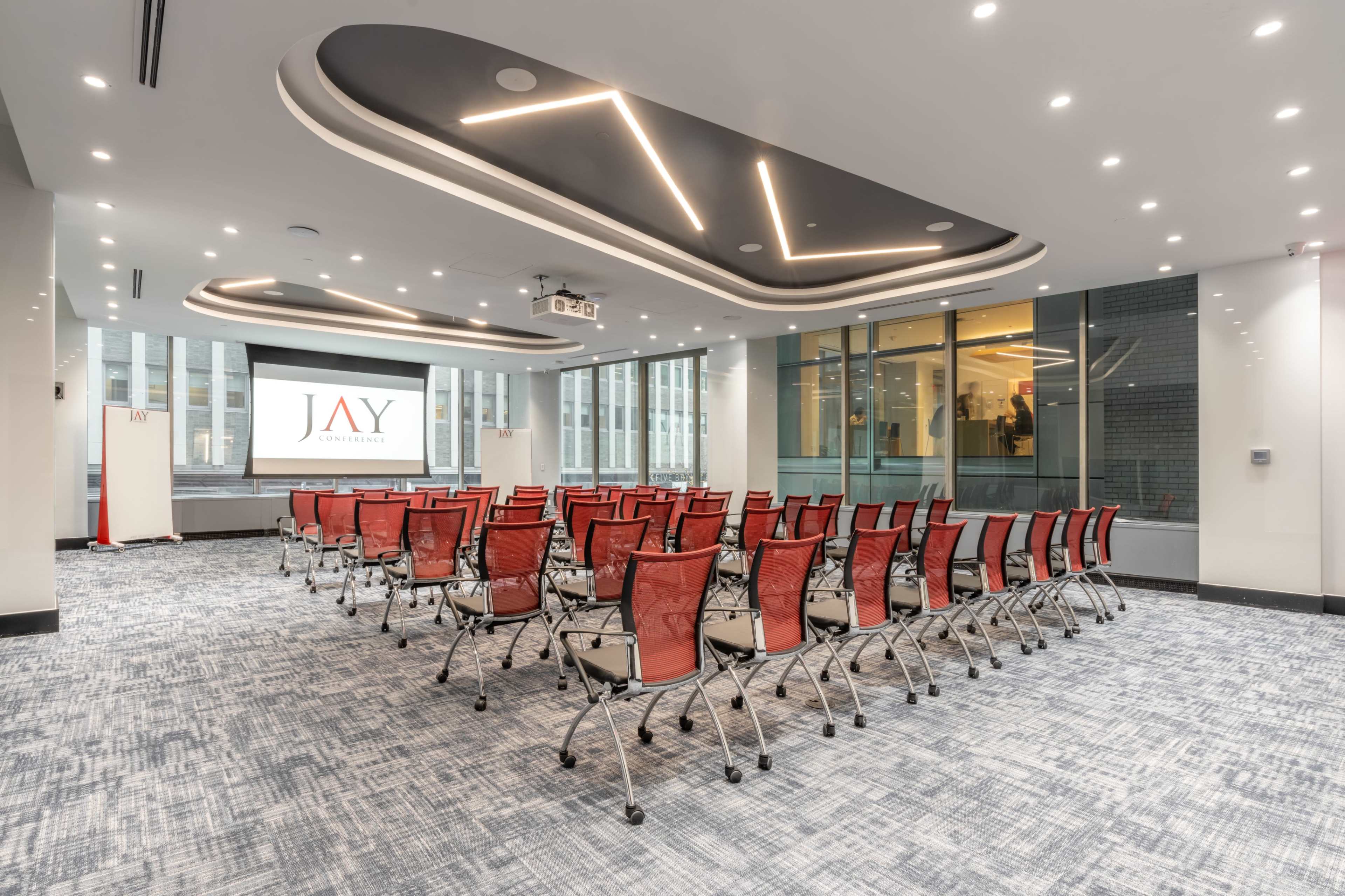 A conference room is set up with rows of red chairs facing a screen displaying the logo "JAY" and has modern architectural lighting.