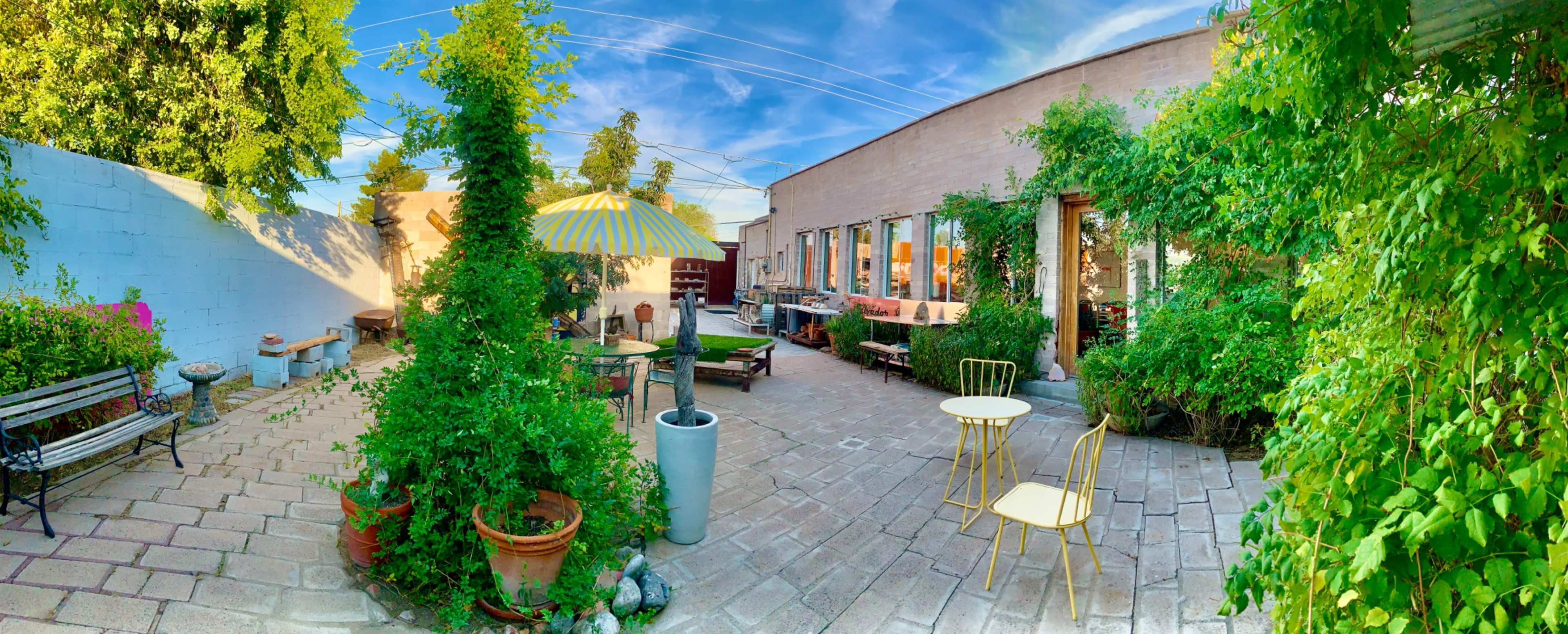 The image shows a garden courtyard with stone pathways, green plants, outdoor seating, and a building in the background.