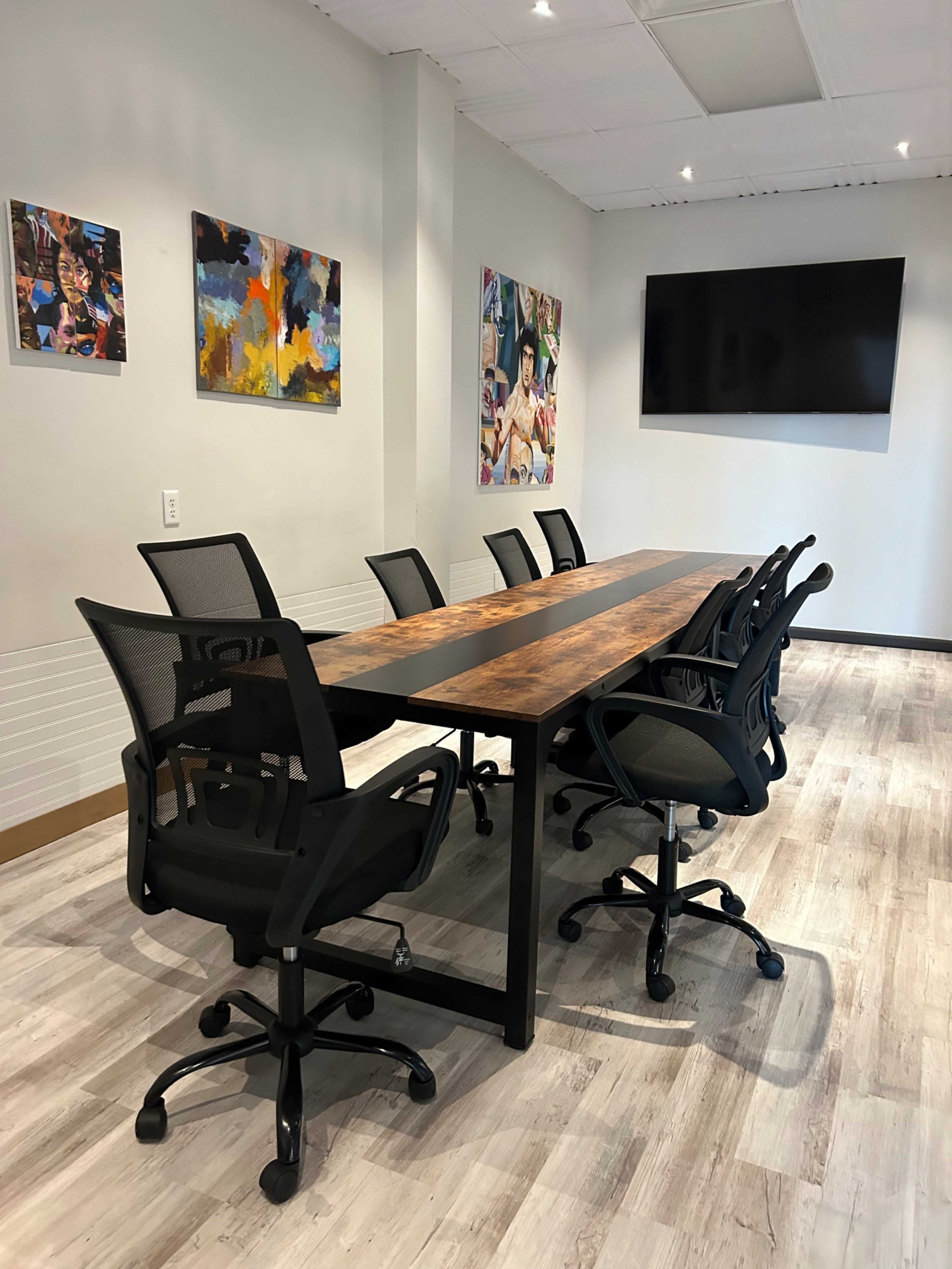 A modern conference room features a large wooden table surrounded by ergonomic chairs, with colorful artwork on the walls and a flat-screen TV mounted at one end.