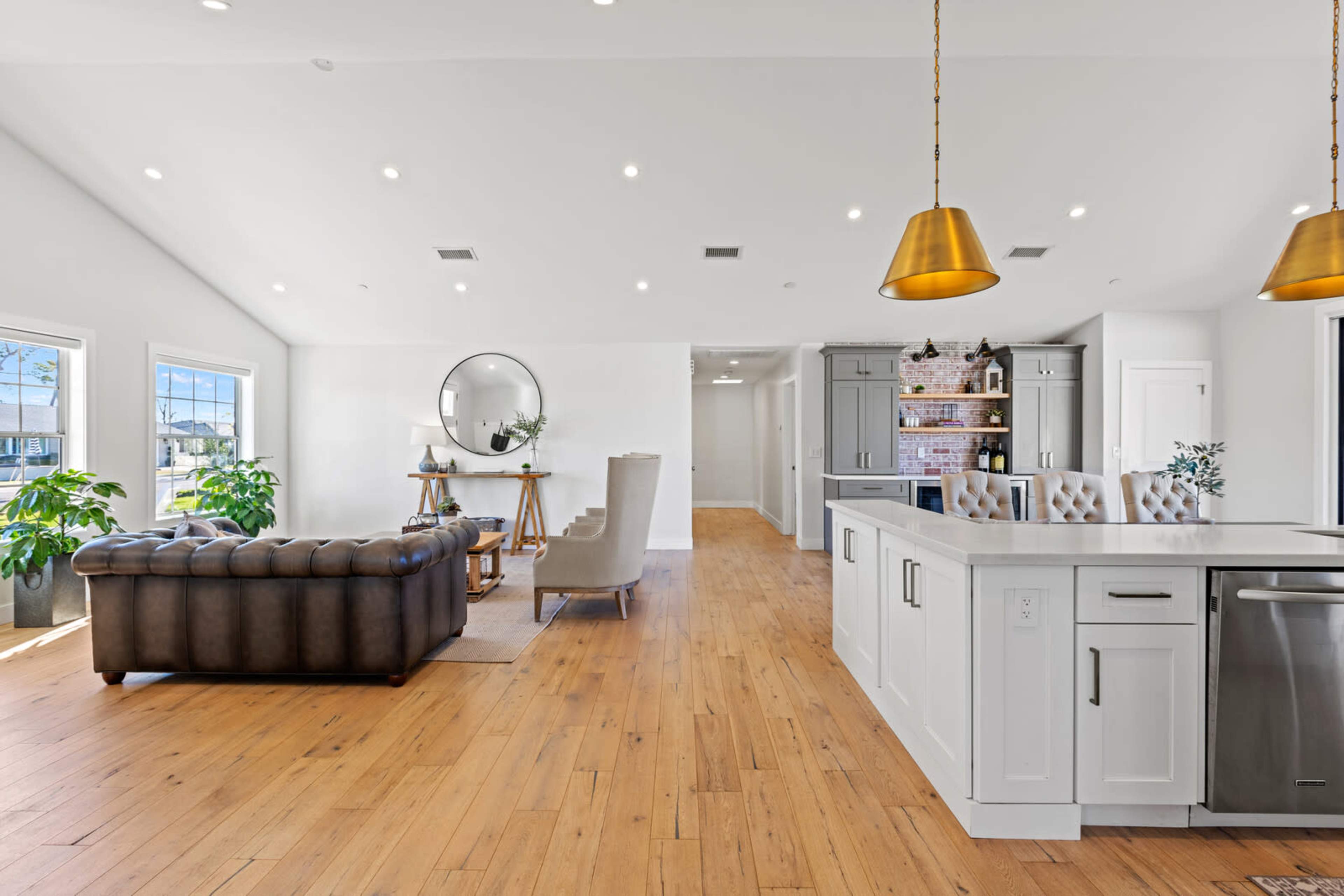 The image shows a spacious, modern living area with a sectional couch, a dining table, and a kitchen featuring white cabinets and a stainless-steel refrigerator.