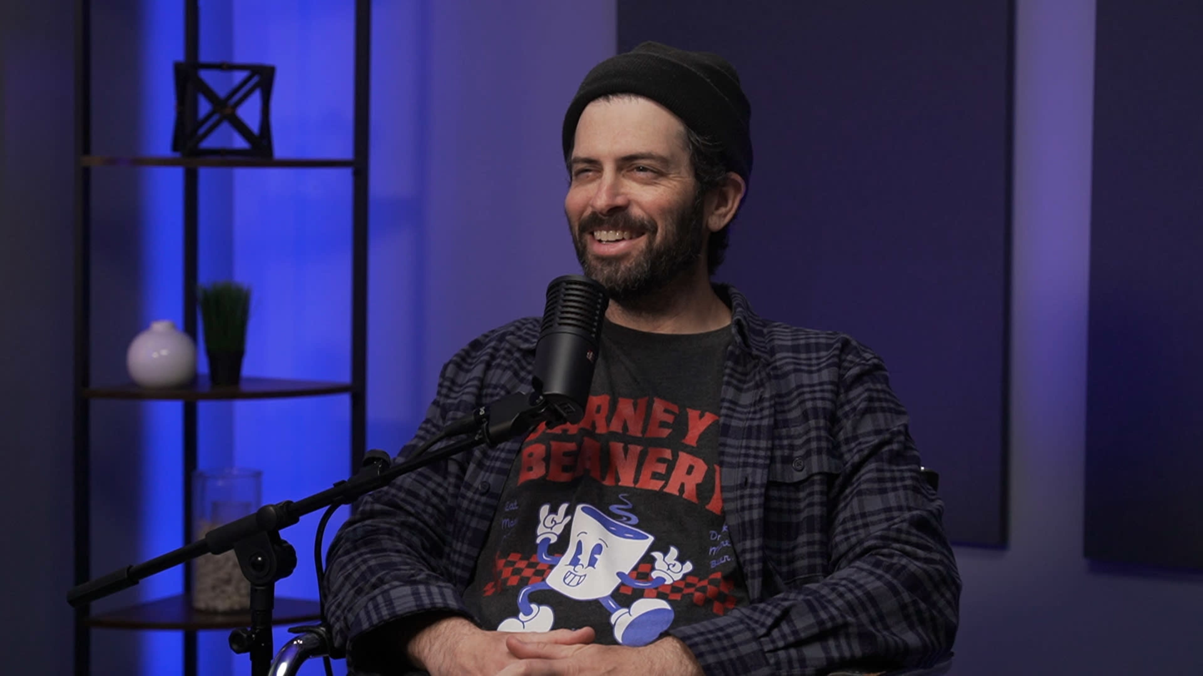 A man with a beard and a beanie smiles while seated at a microphone in a studio setting with blue lighting.