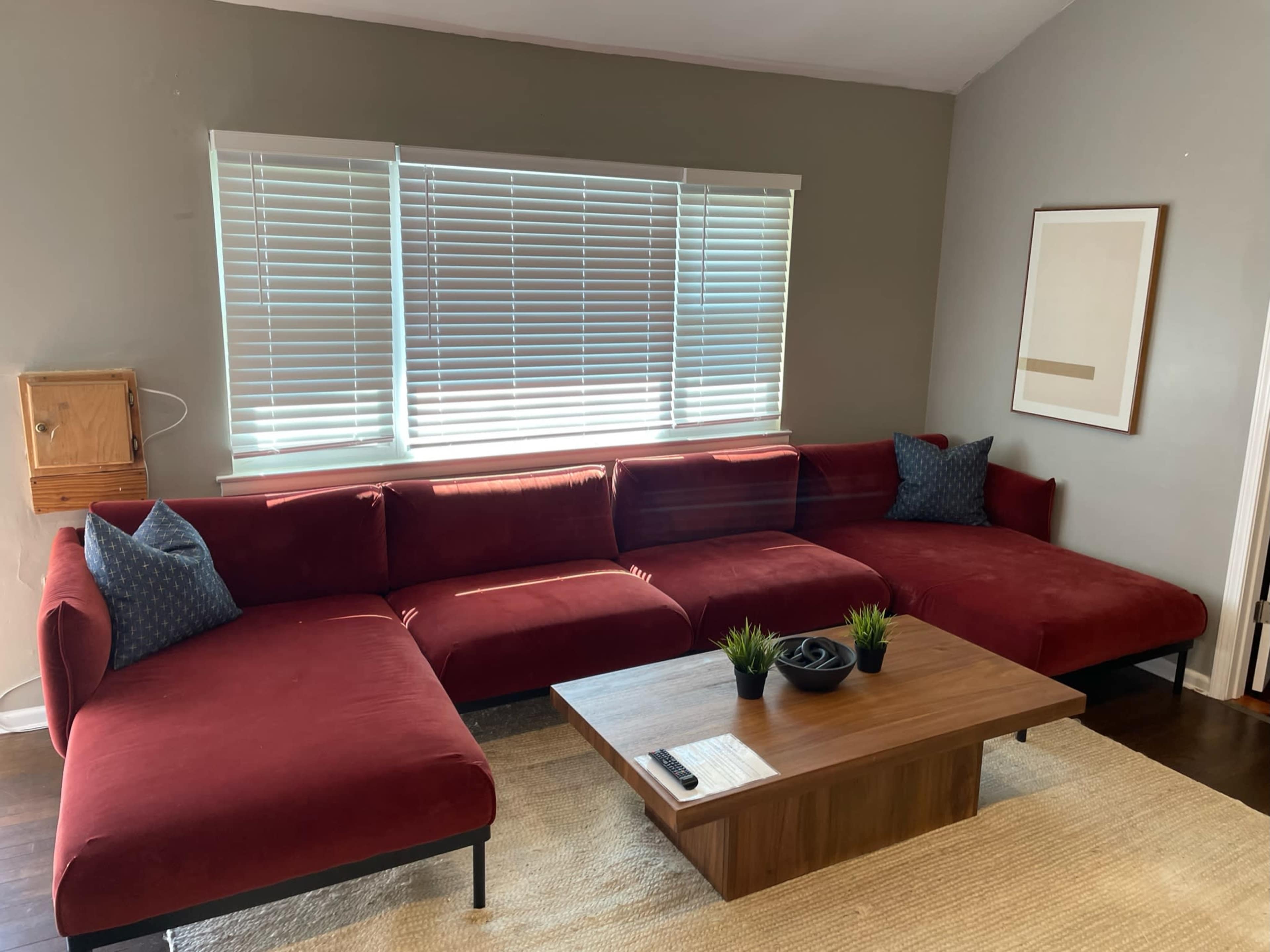 A red sectional sofa with blue accent pillows accompanies a wooden coffee table in a room featuring large windows covered with white blinds.