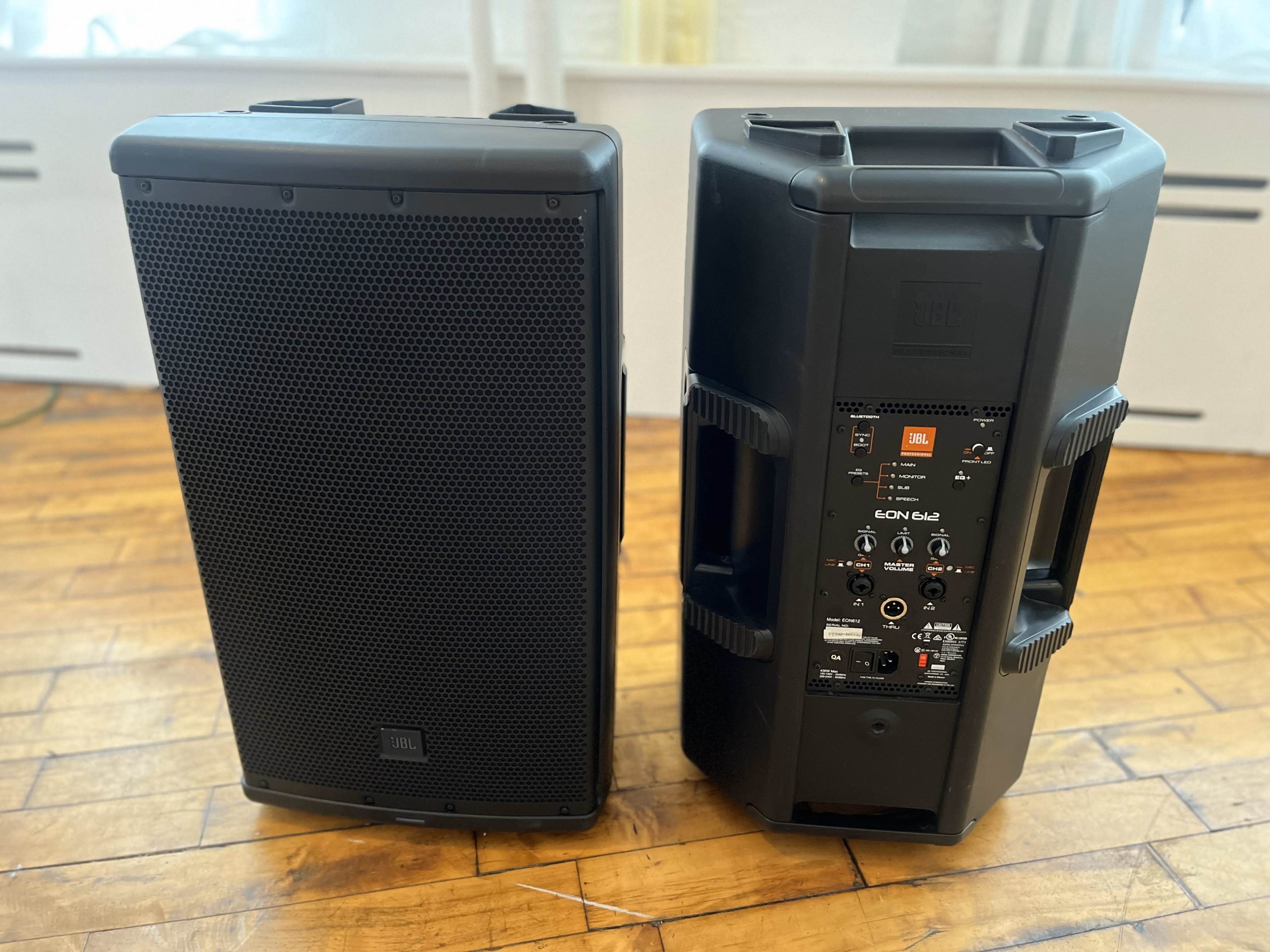 The image shows two JBL EON610 loudspeakers positioned on a wooden floor, with one facing forward and the other showing its rear panel controls.