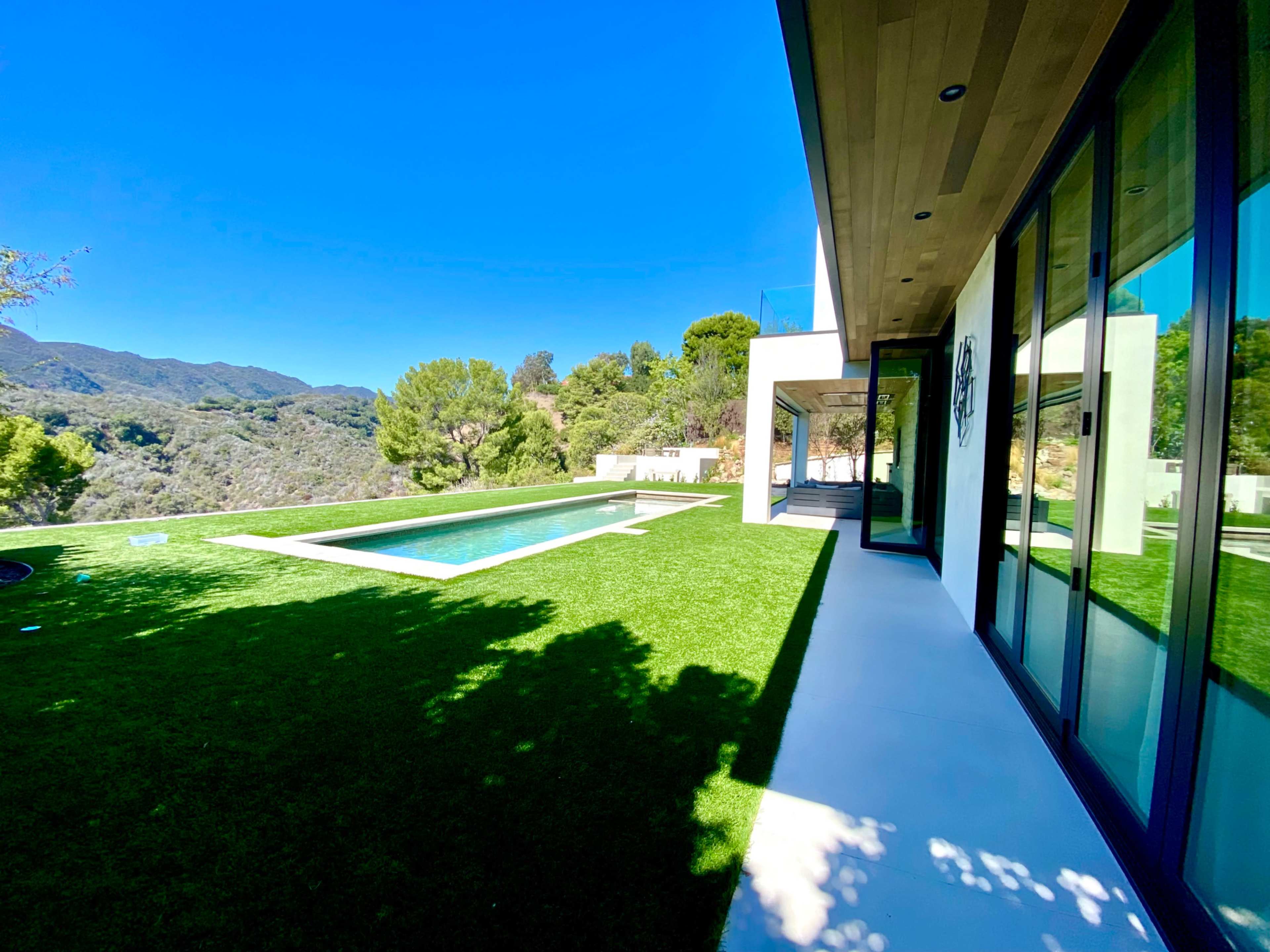 A modern house with large windows overlooks a green lawn and a swimming pool, set against a backdrop of hills and clear blue skies.
