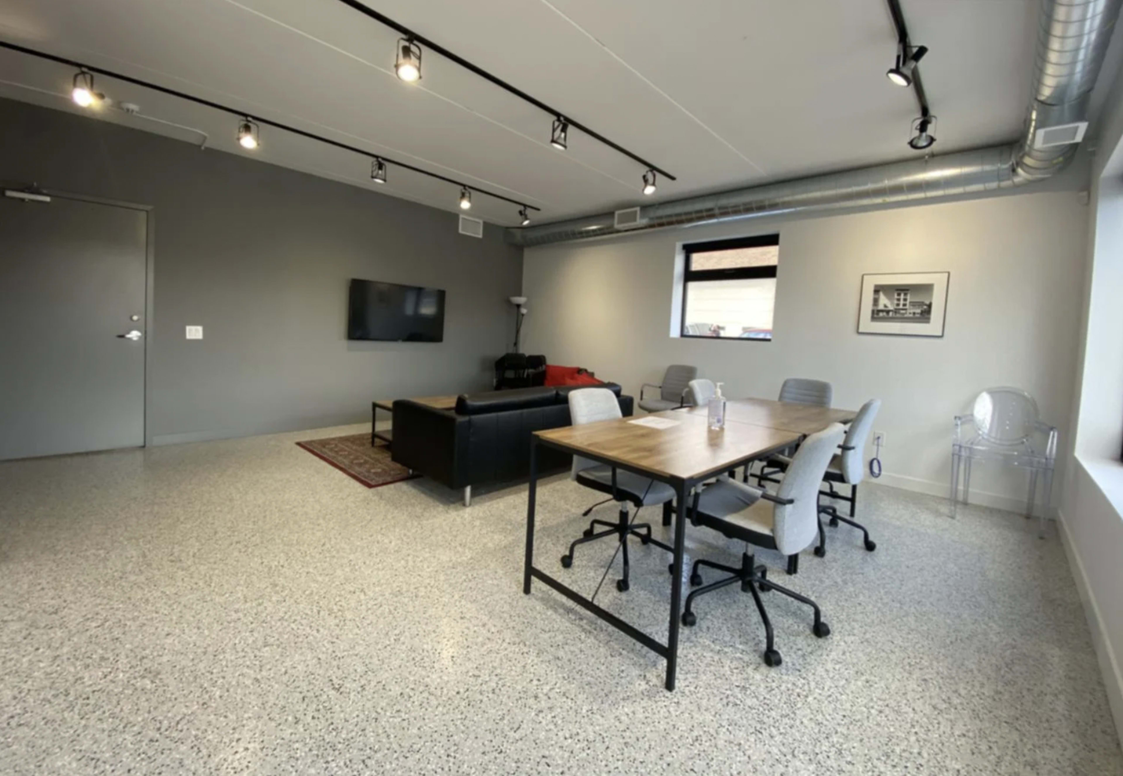 A modern office space featuring a seating area with a black sofa and a wall-mounted TV, along with a conference table surrounded by chairs.