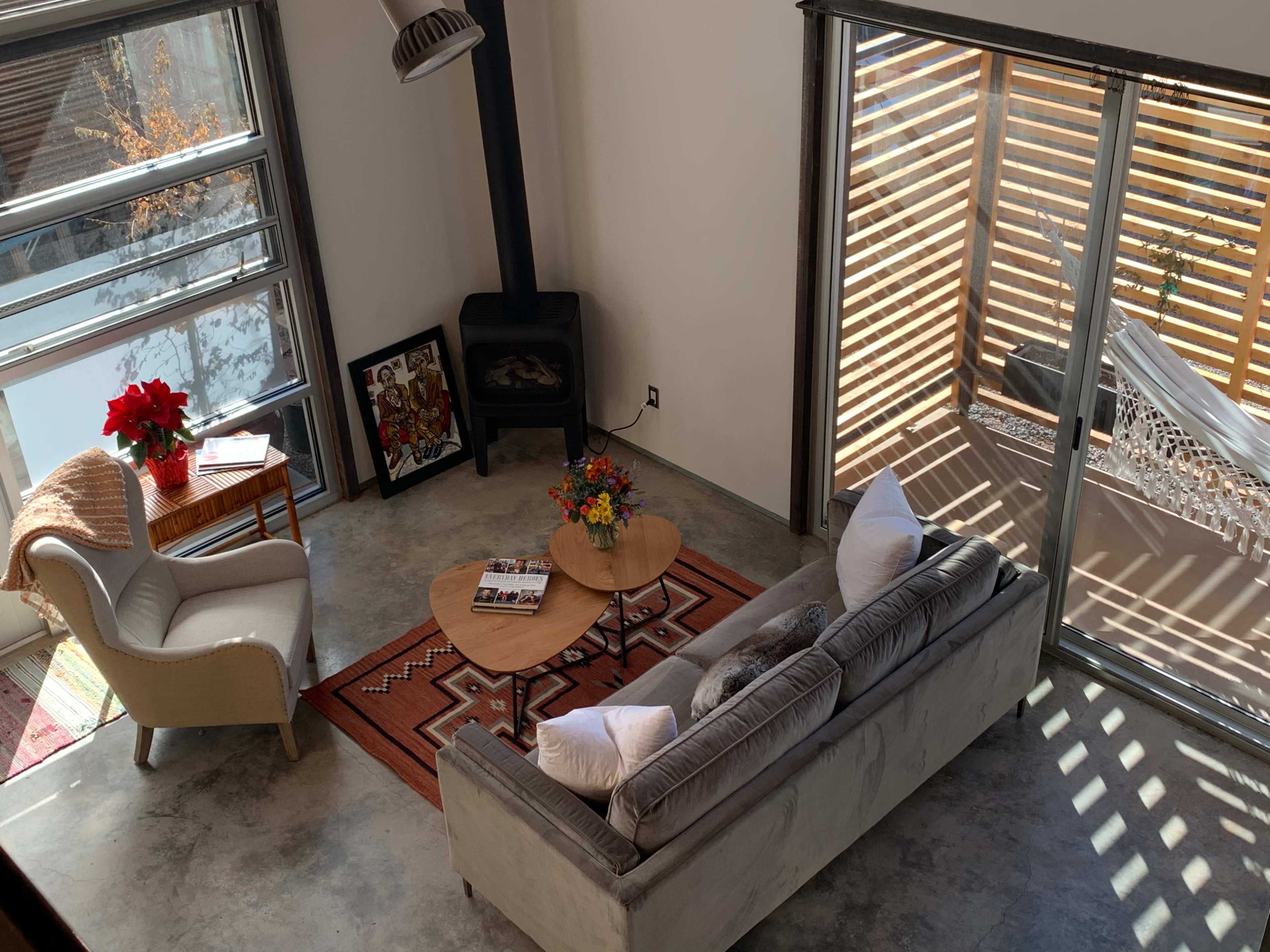 A living room features a gray sofa, a round wooden coffee table, a small side table with a plant, and large glass doors that open to an outdoor space with shade slats.