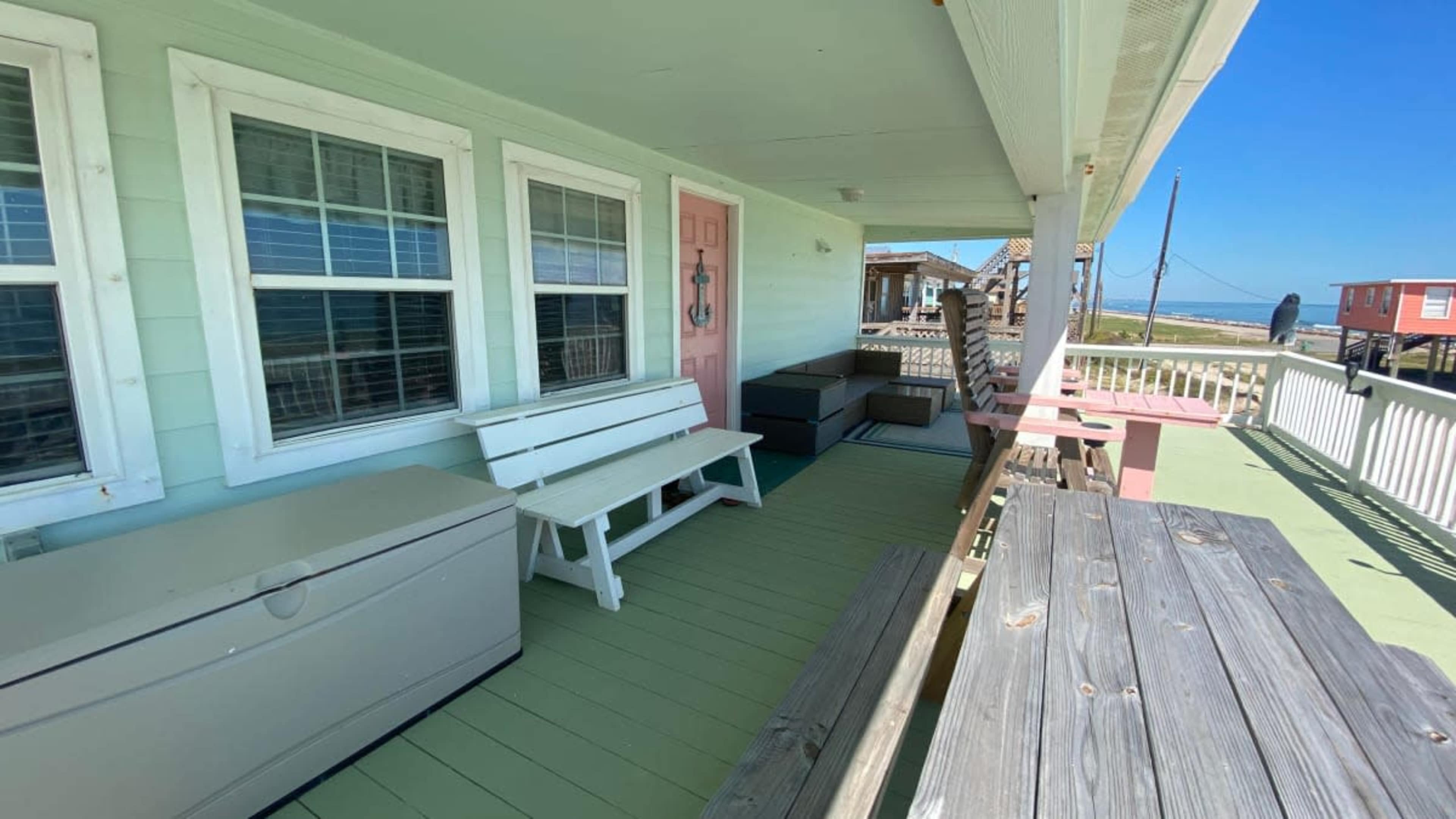 The image shows a porch with pale green flooring, white benches, and a pink door, overlooking a coastal view.